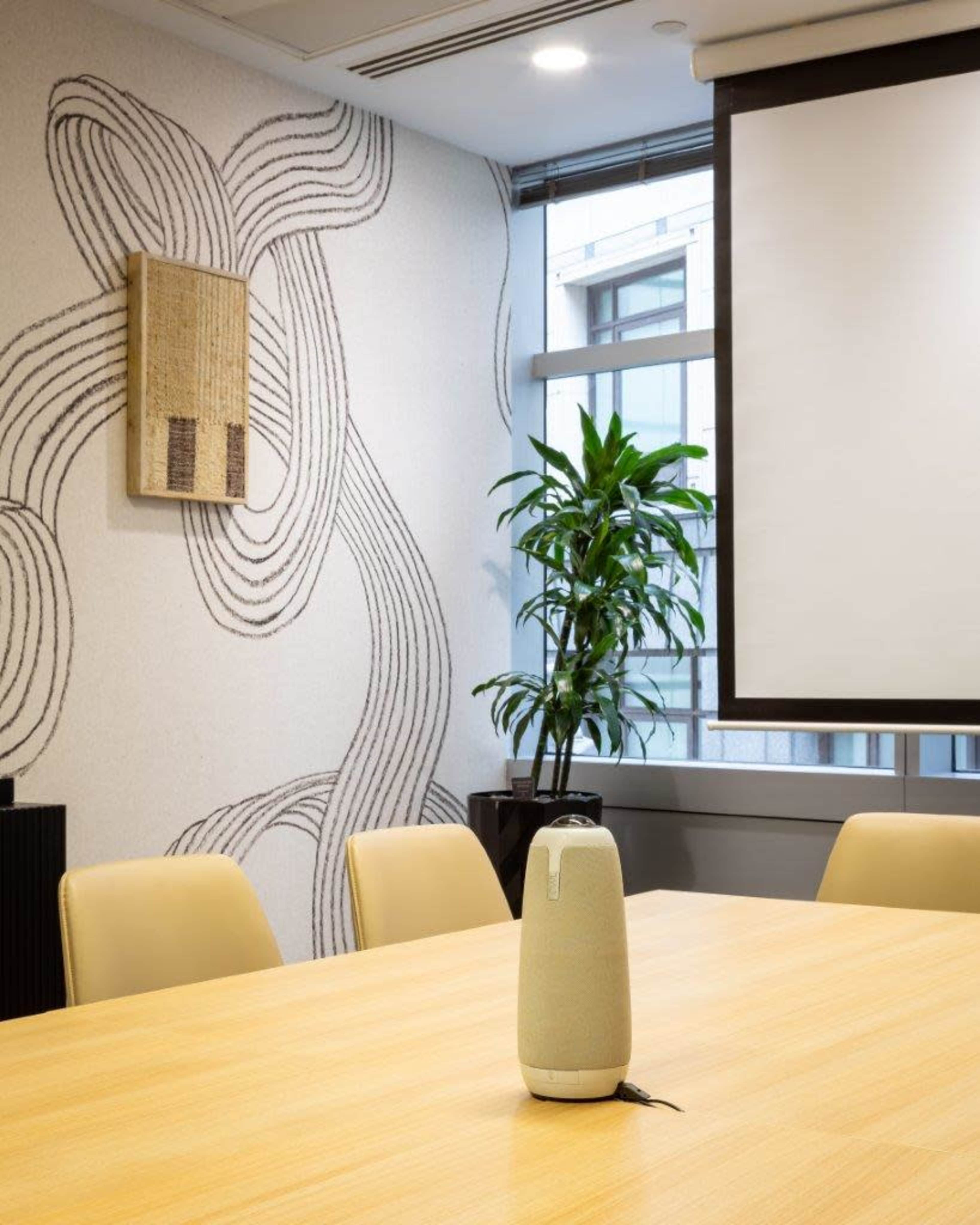 The image shows a modern conference room with a round wooden table, a decorative wall with abstract patterns, and a potted plant near a large window.