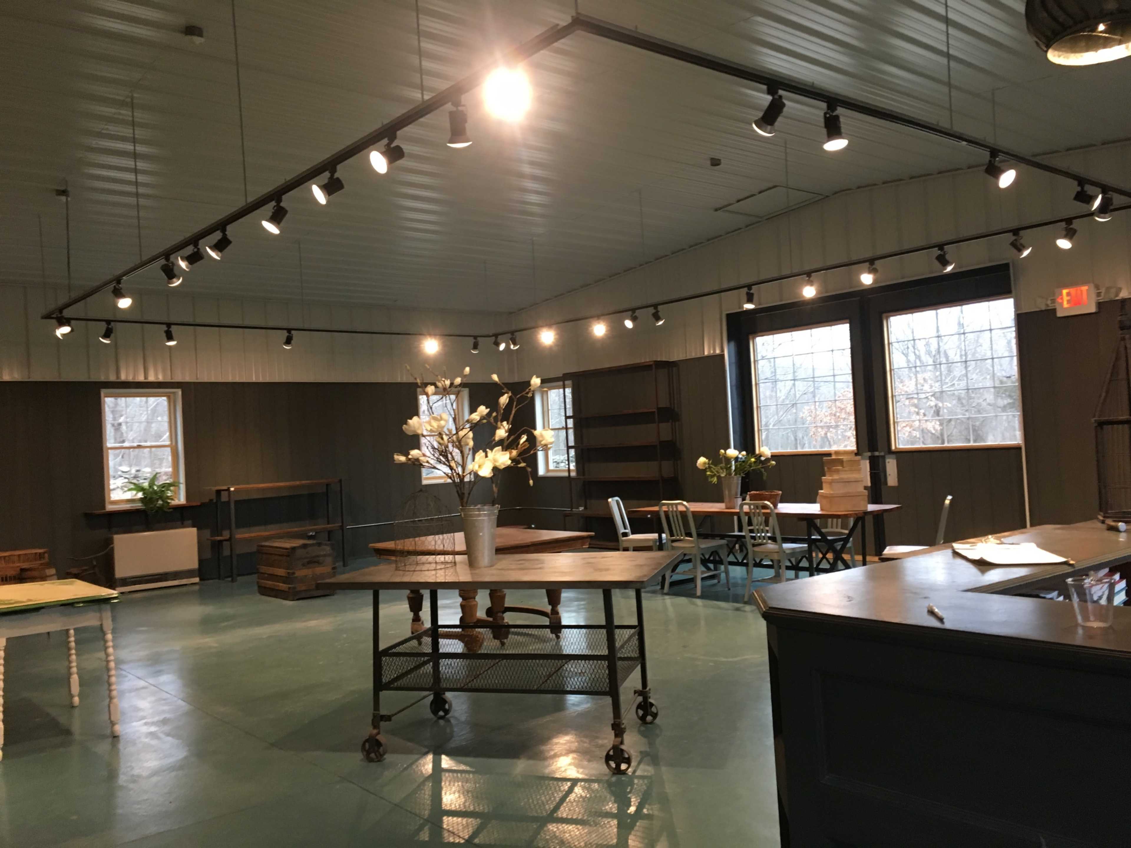 An empty interior space with hardwood floors, featuring several tables, shelves, and large windows illuminating the area.