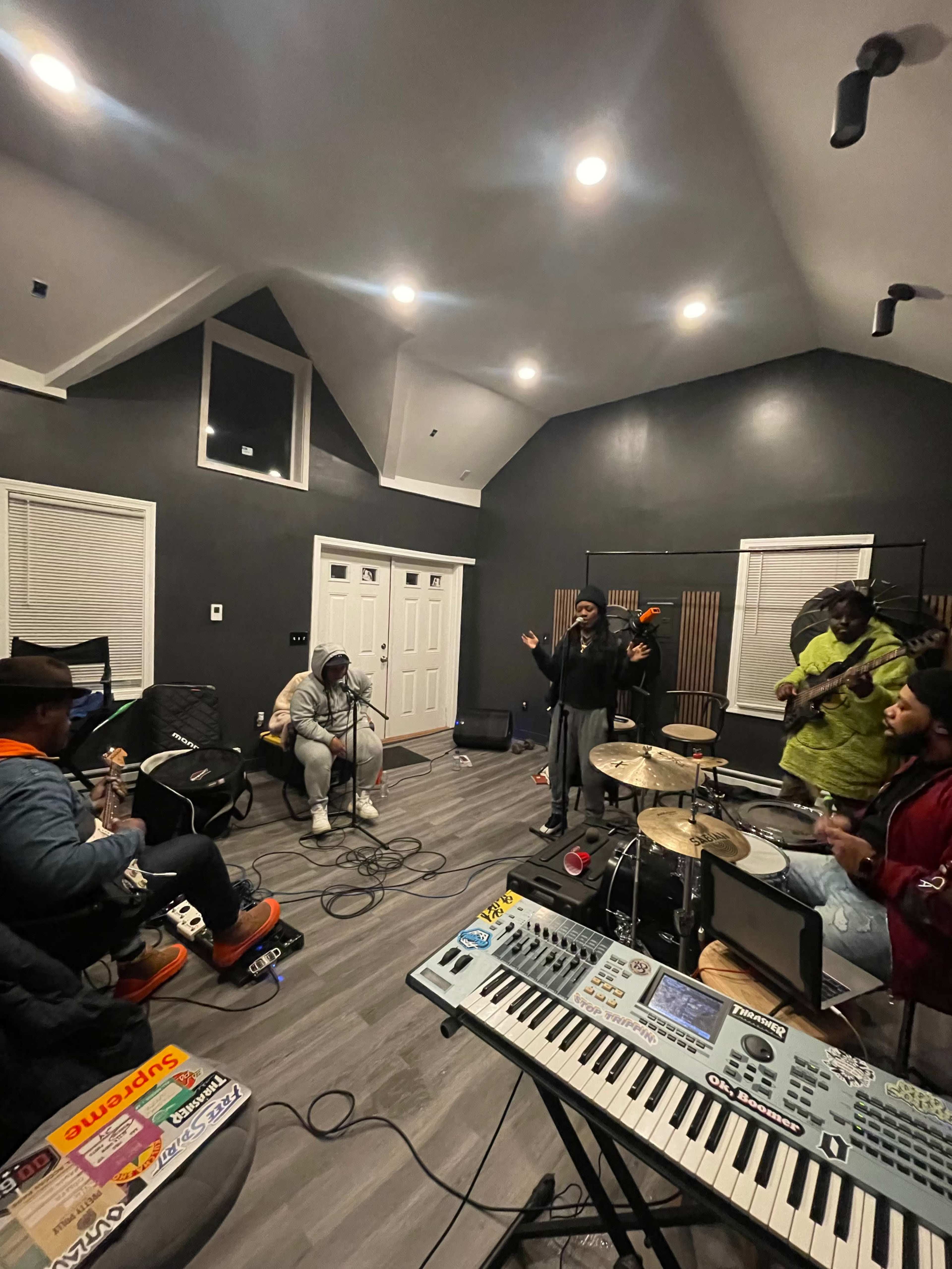 A group of musicians plays instruments and sings in a spacious recording studio with modern lighting and sound equipment.