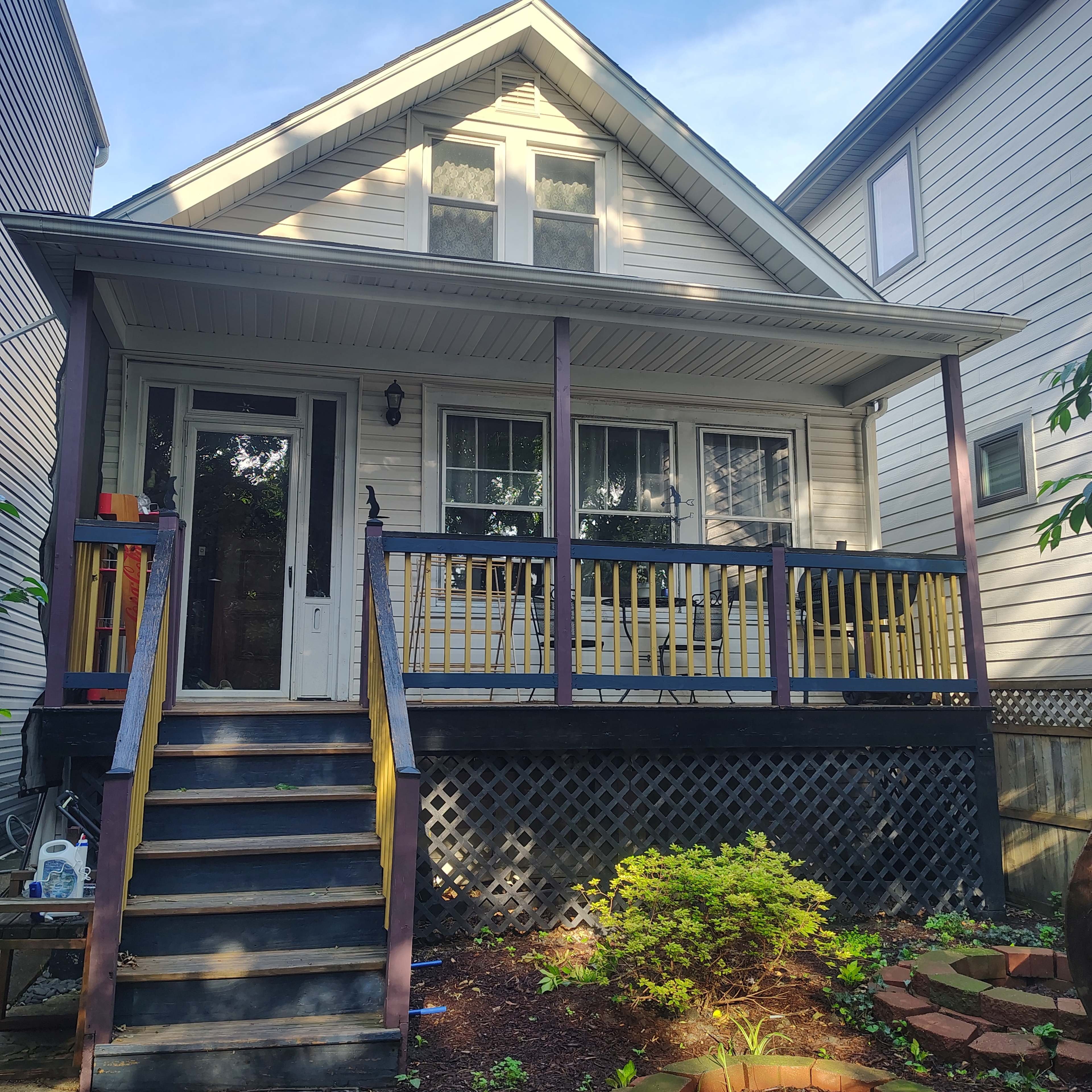 The image shows a two-story house with a front porch and stairs leading up to the entrance, surrounded by landscaping and adjacent buildings.