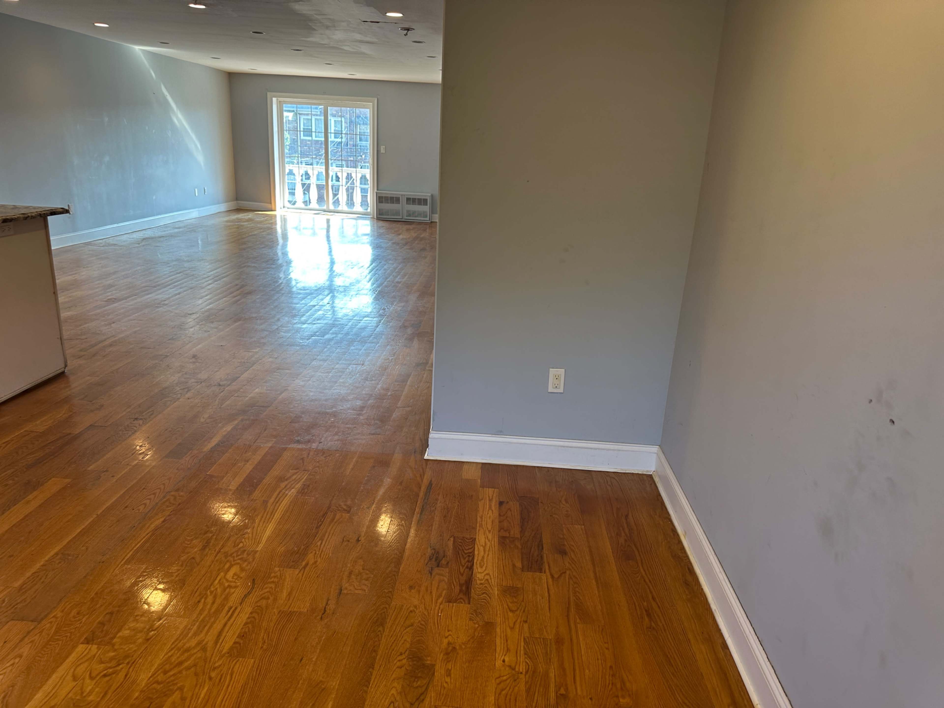 The image shows a spacious room with polished hardwood floors, leading towards a doorway that opens to a balcony.