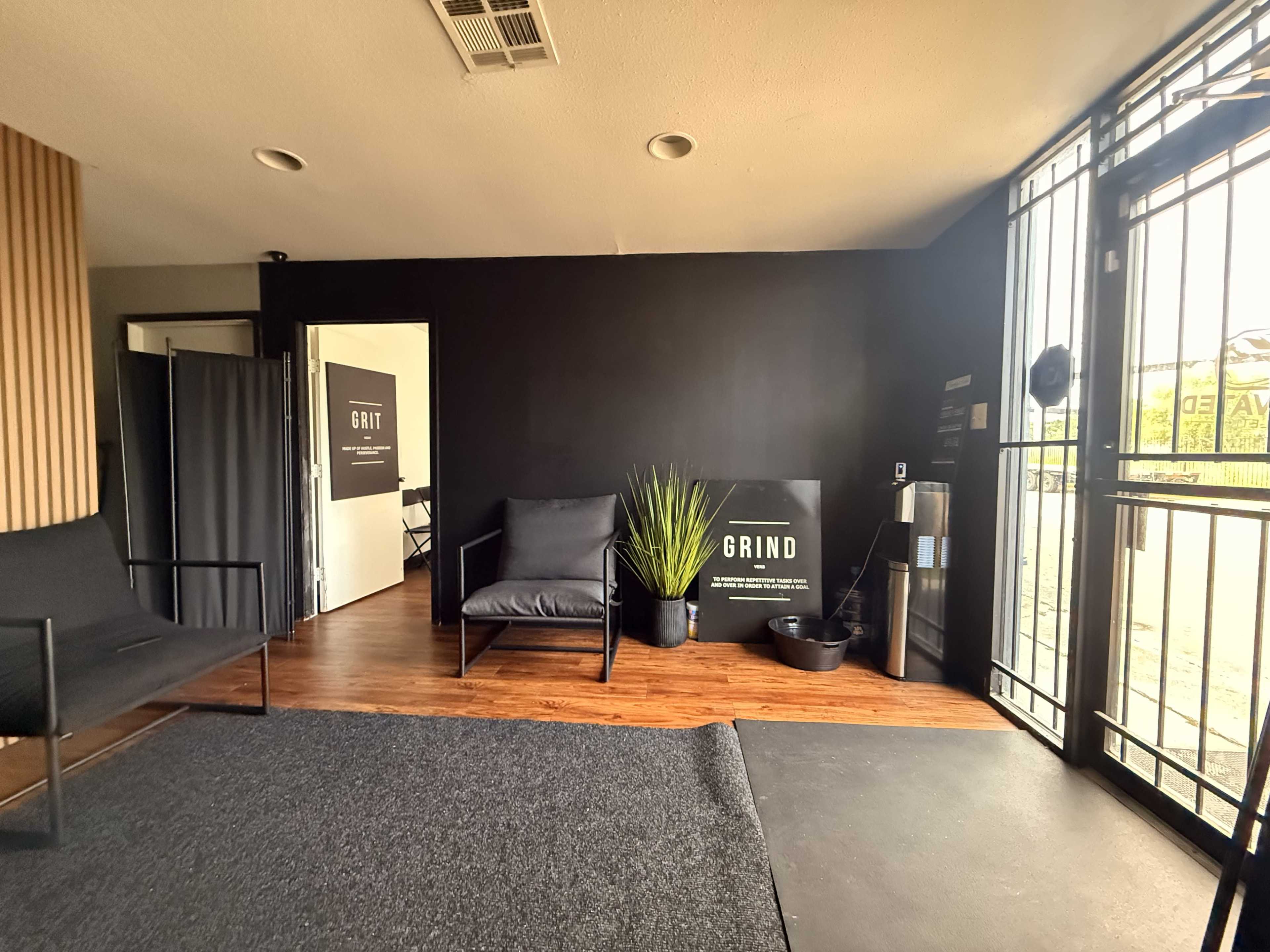 The image shows a modern interior space with black walls, wooden flooring, and minimalist furniture, including two chairs and a plant.