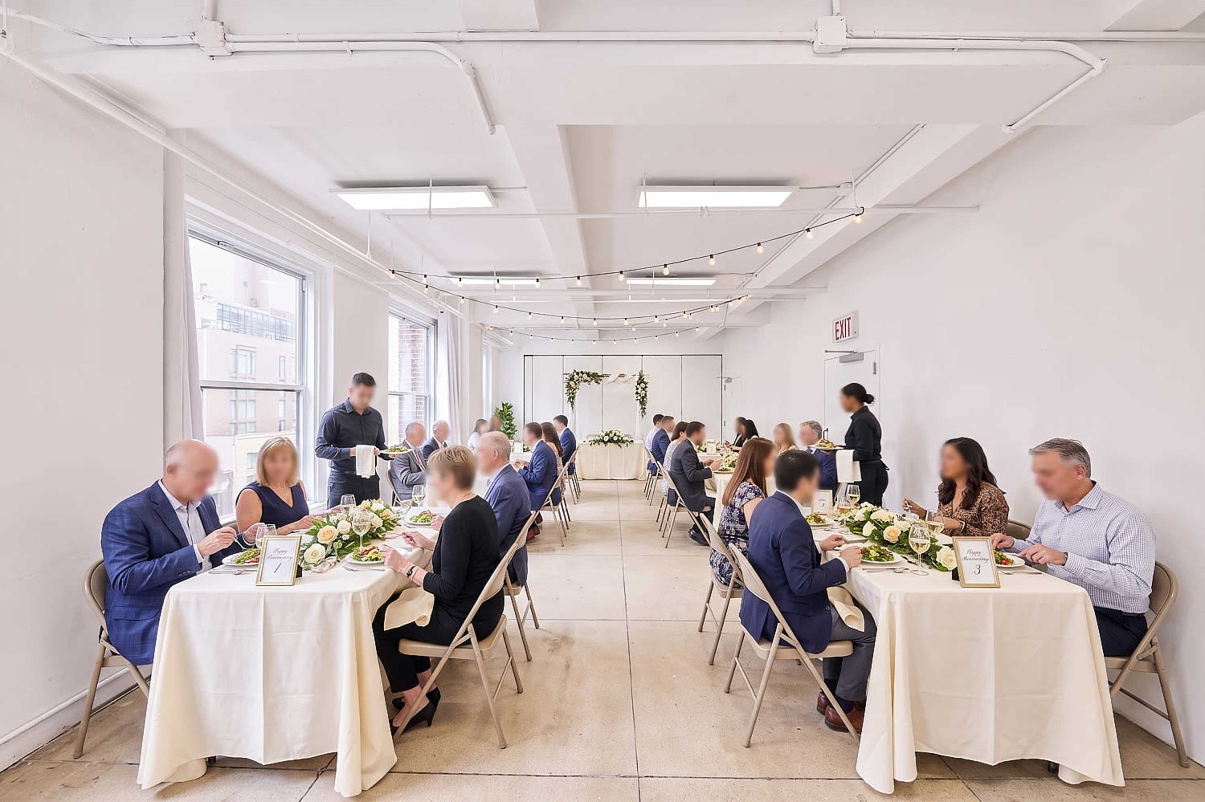A group of people is seated at tables in a large, well-lit room, dining amid floral centerpieces and string lights.
