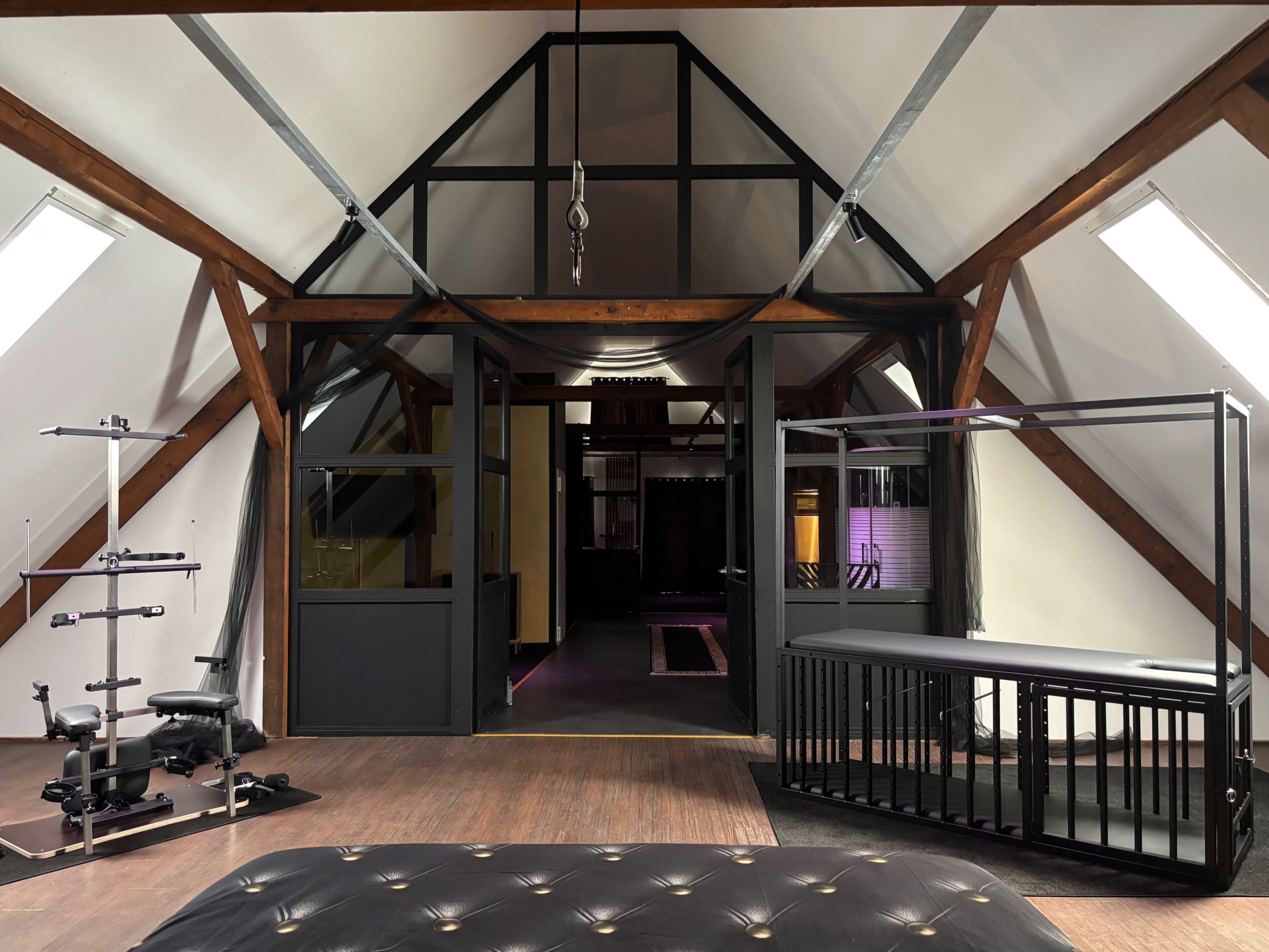 The image shows an attic space featuring exposed wooden beams, gym equipment on the left, and a large open area that leads to another room in the background.