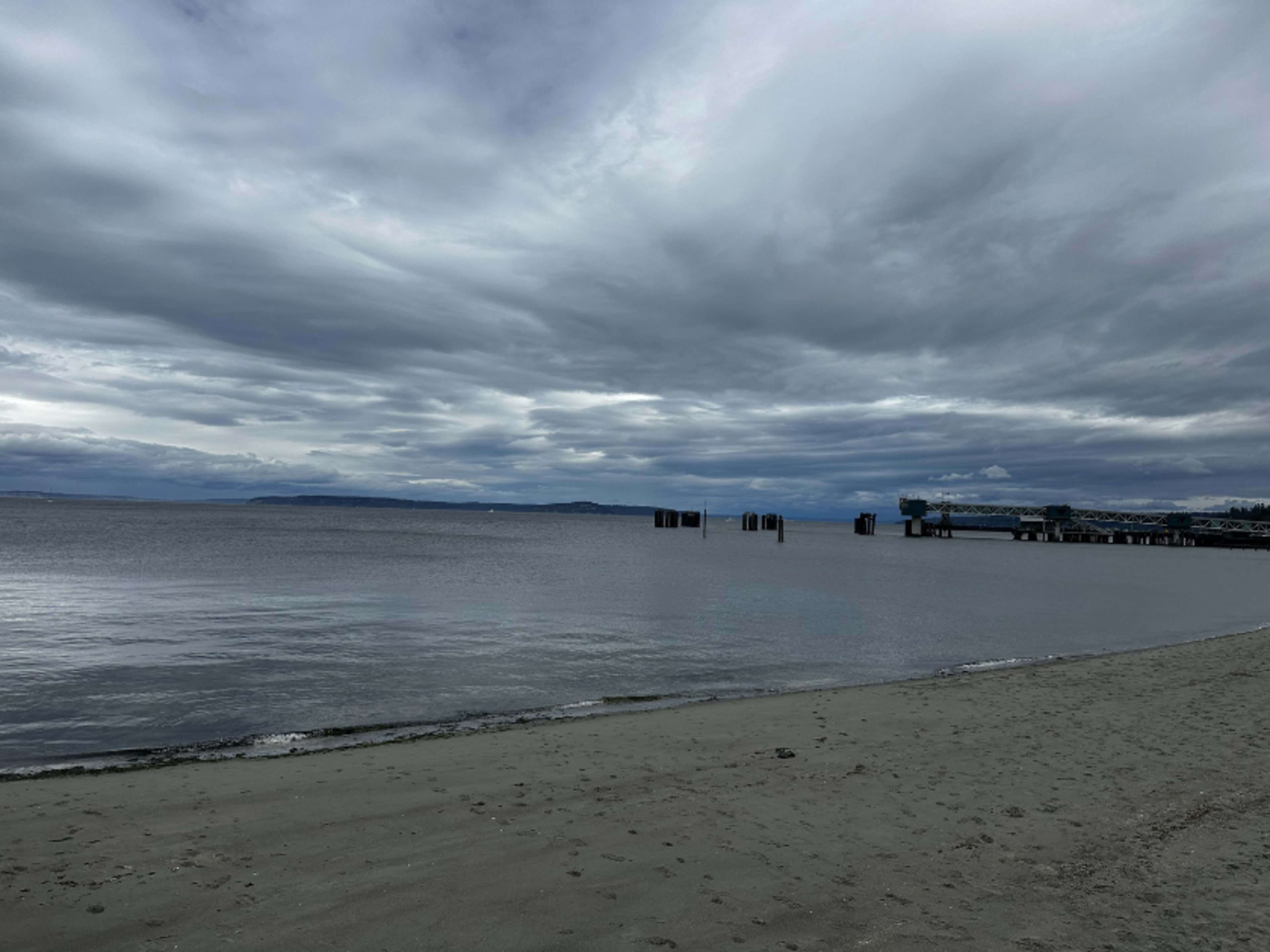 The image shows a calm beach with a sandy shore, gray clouds overhead, and a distant row of wooden posts extending into the water.