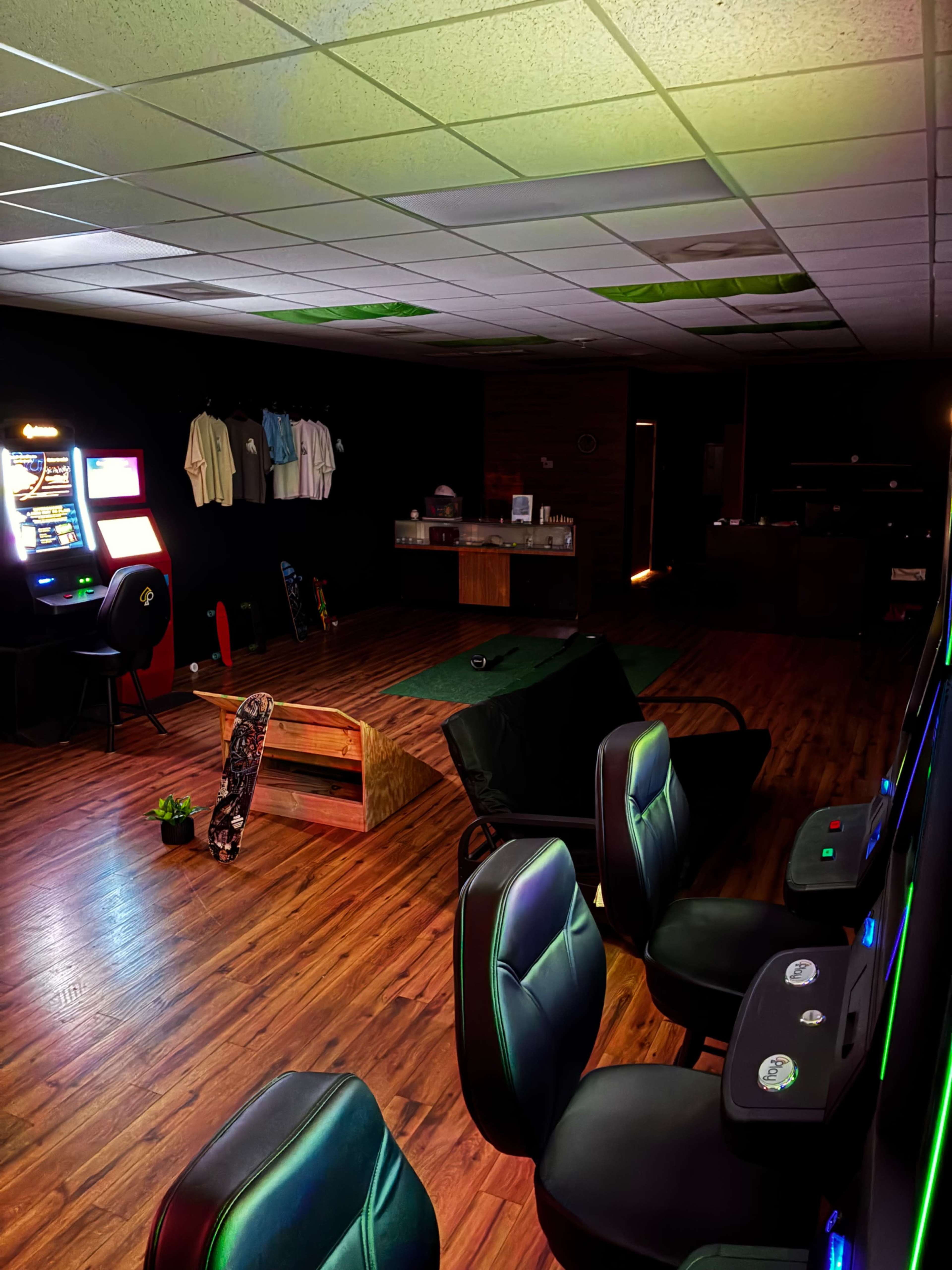 The image shows an indoor gaming lounge with arcade machines, a skateboard ramp, and chairs arranged in a darkened room with wooden flooring.