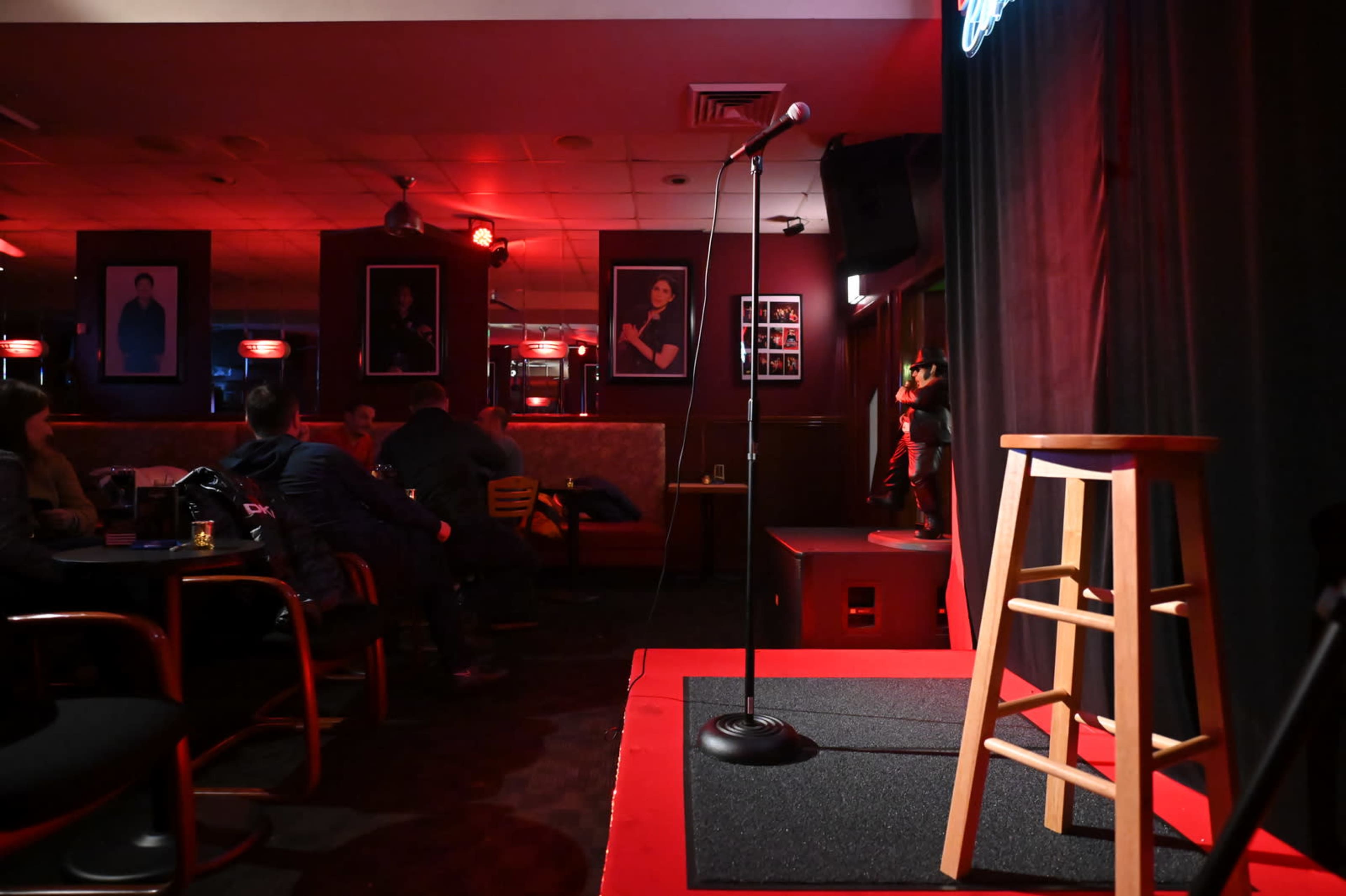 A microphone stands on a stool in a dimly lit comedy club, with spectators seated at tables in the background.