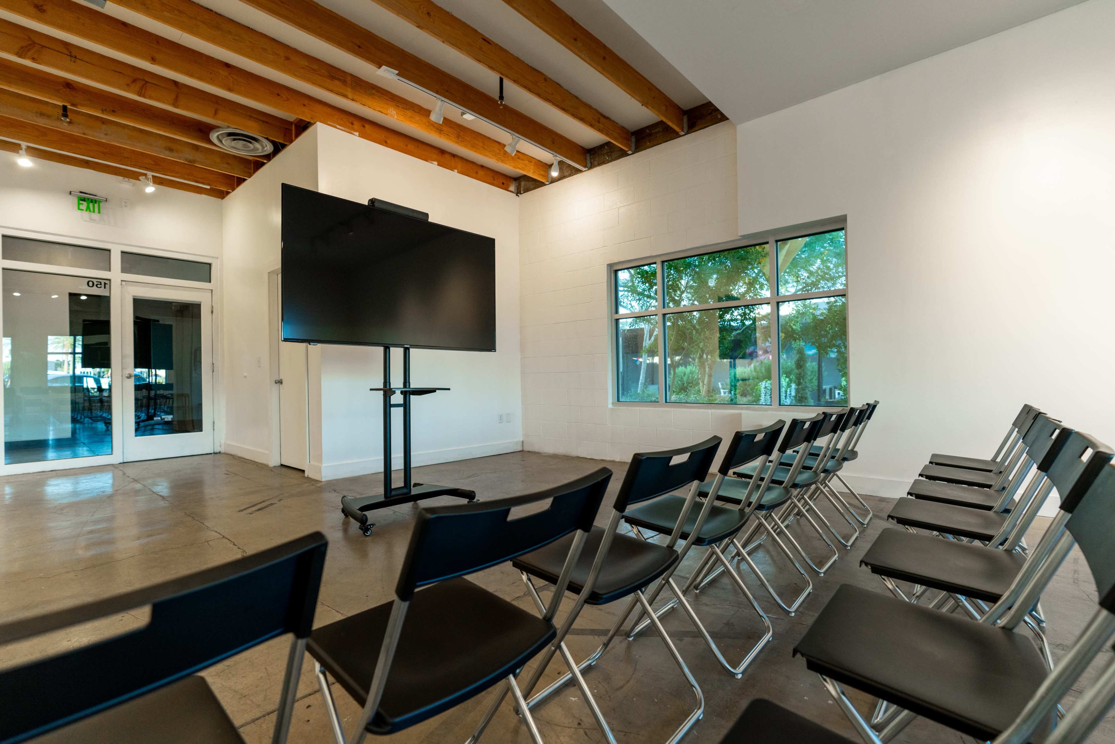 A sparse meeting room with rows of black chairs facing a large screen on a movable stand, near a window and an entrance door.