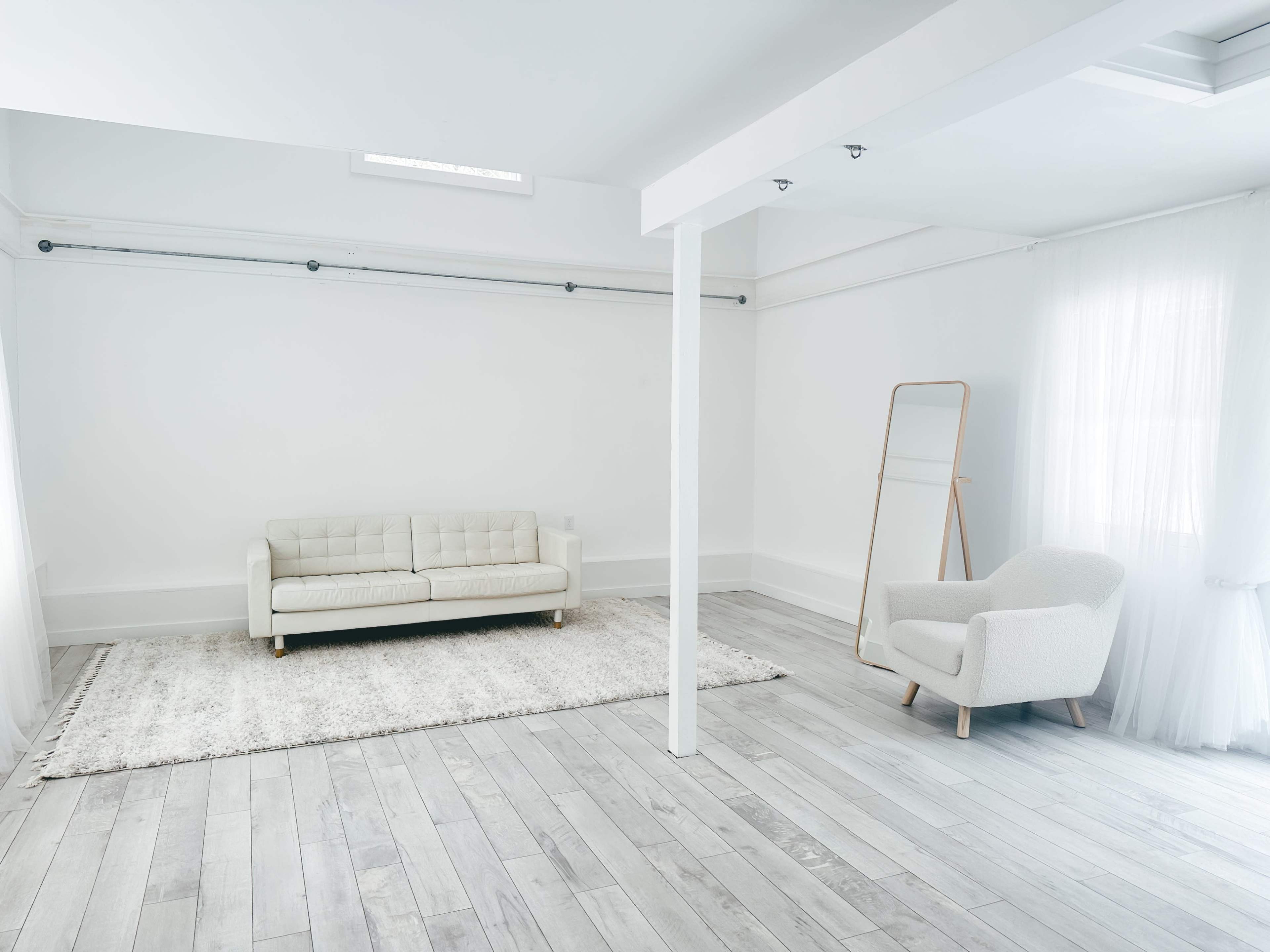A minimalist room with a light-colored wooden floor, a white sofa, a rug, and a mirror leaning against the wall.