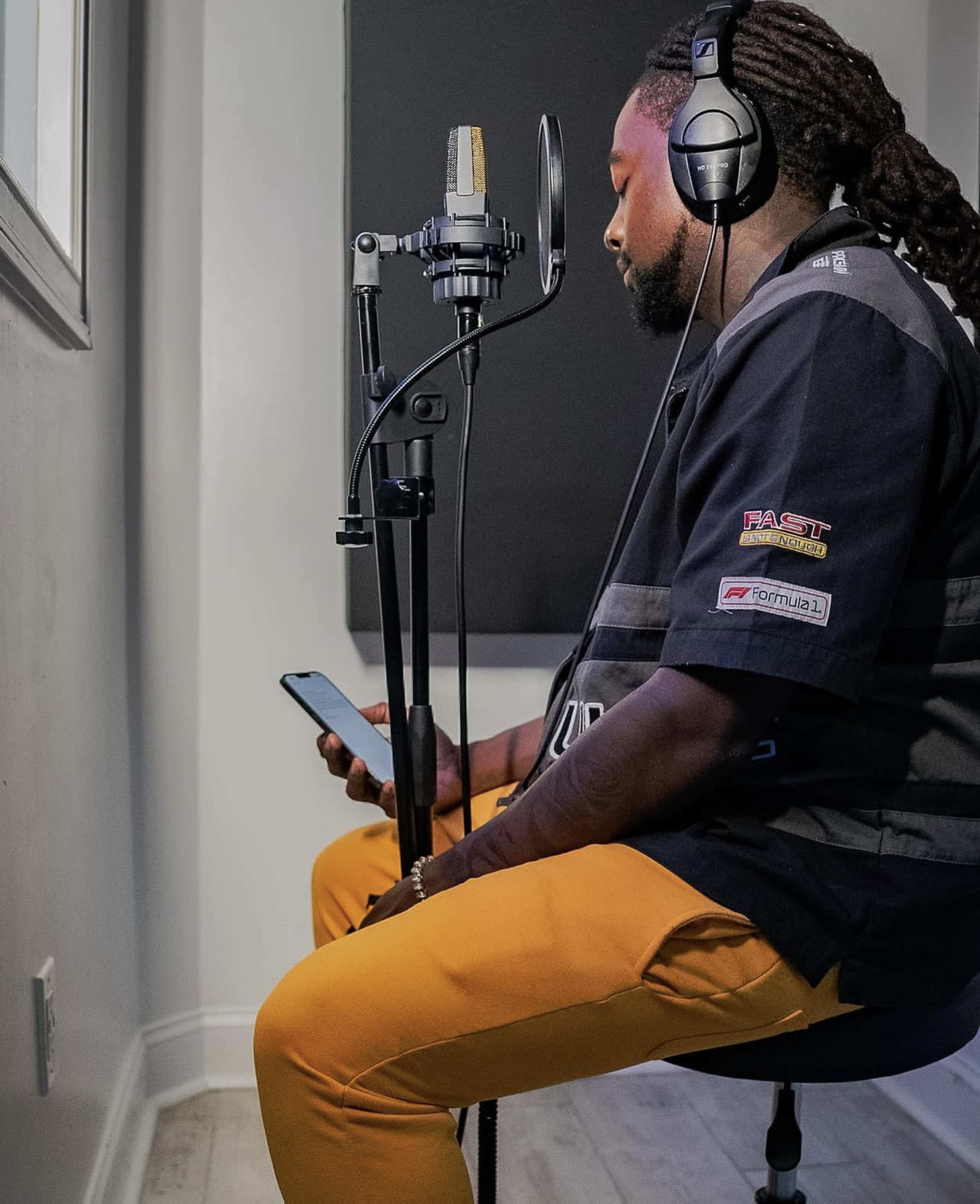 A person wearing headphones is seated in a recording booth, looking at their phone while positioned near a microphone on a stand.