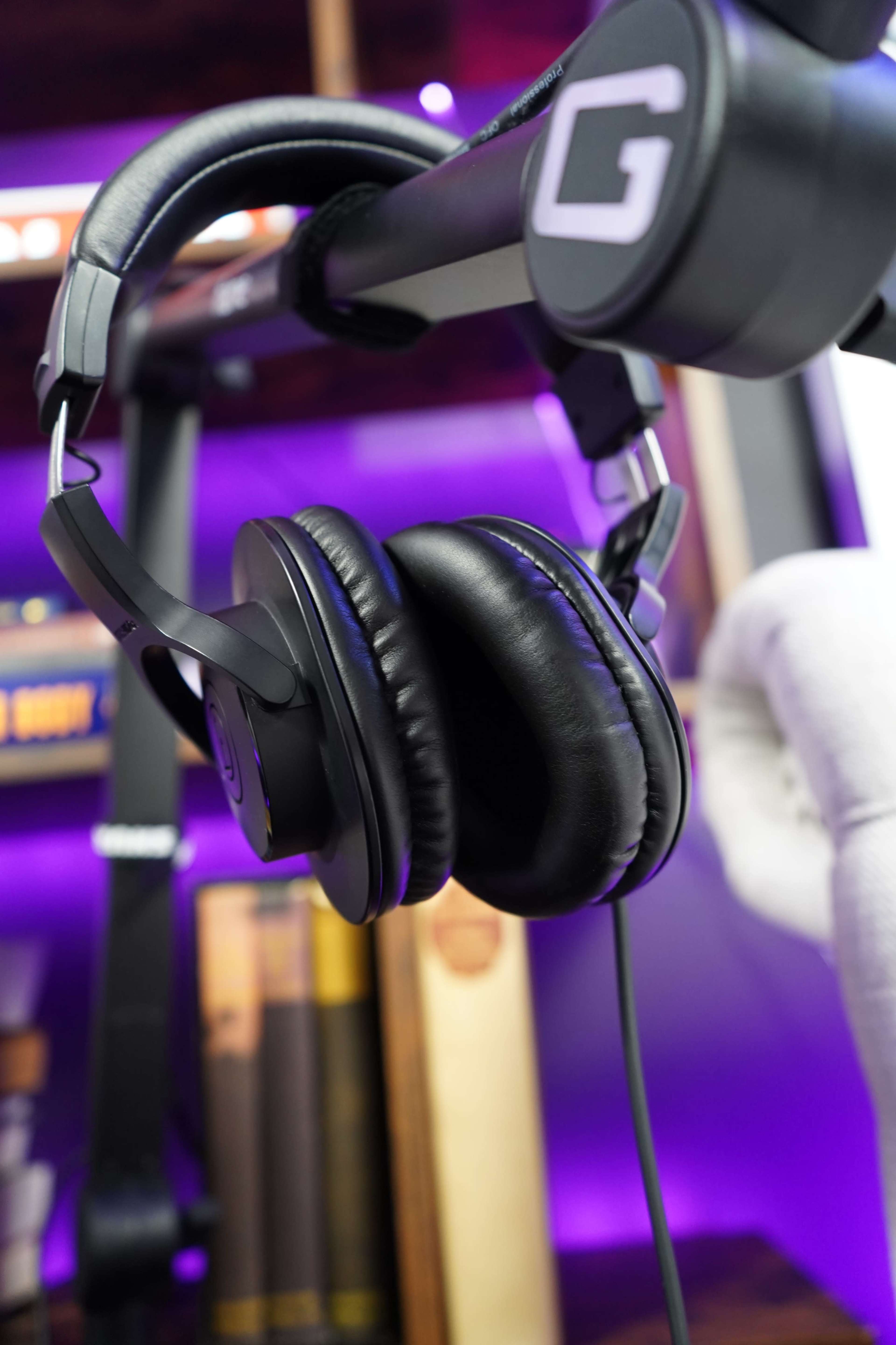 A pair of black headphones hangs from a sleek stand against a backdrop of purple lighting and books.