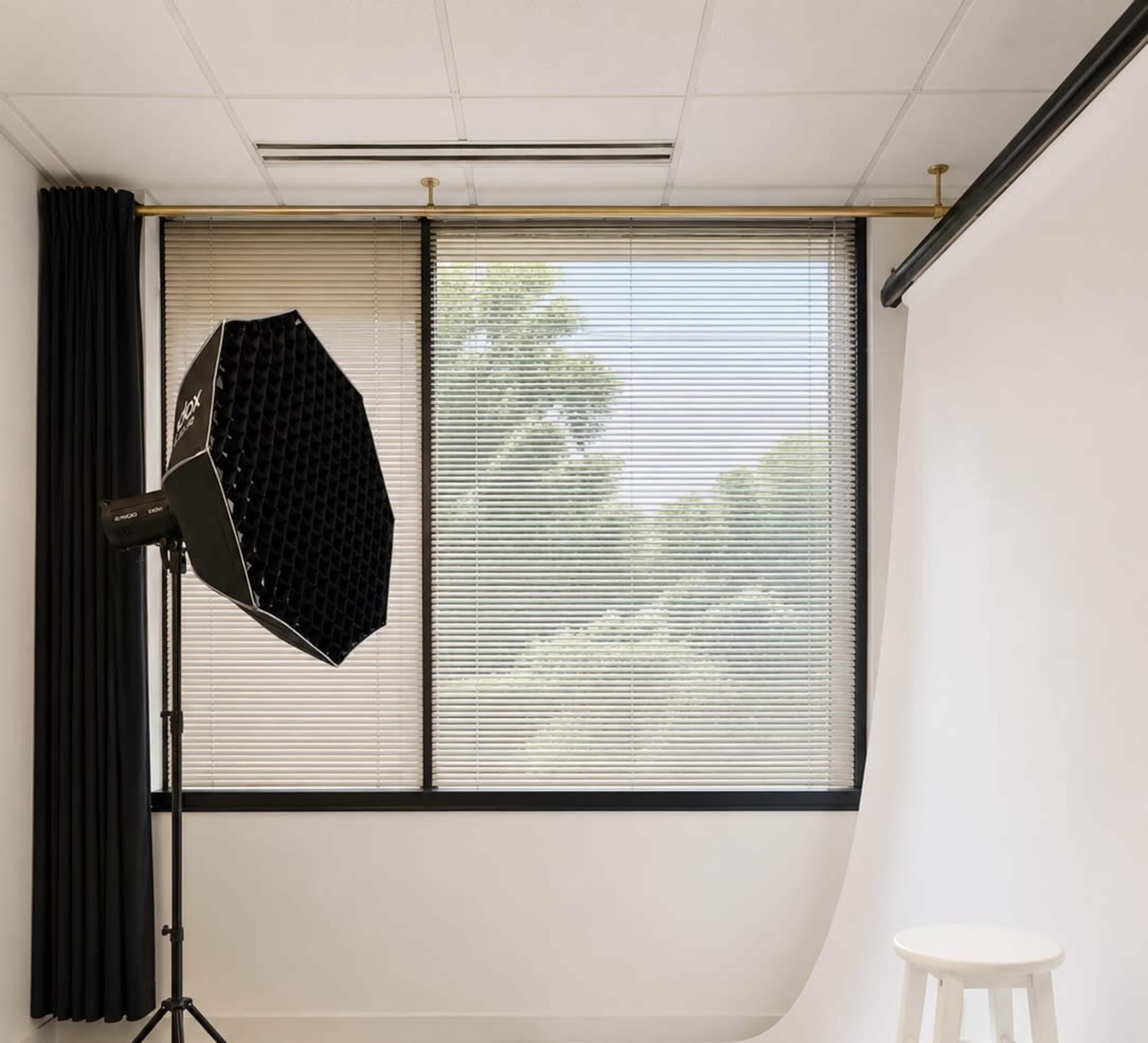 A photography studio features a light modifier on a stand, a white backdrop, and large windows with blinds letting in natural light.