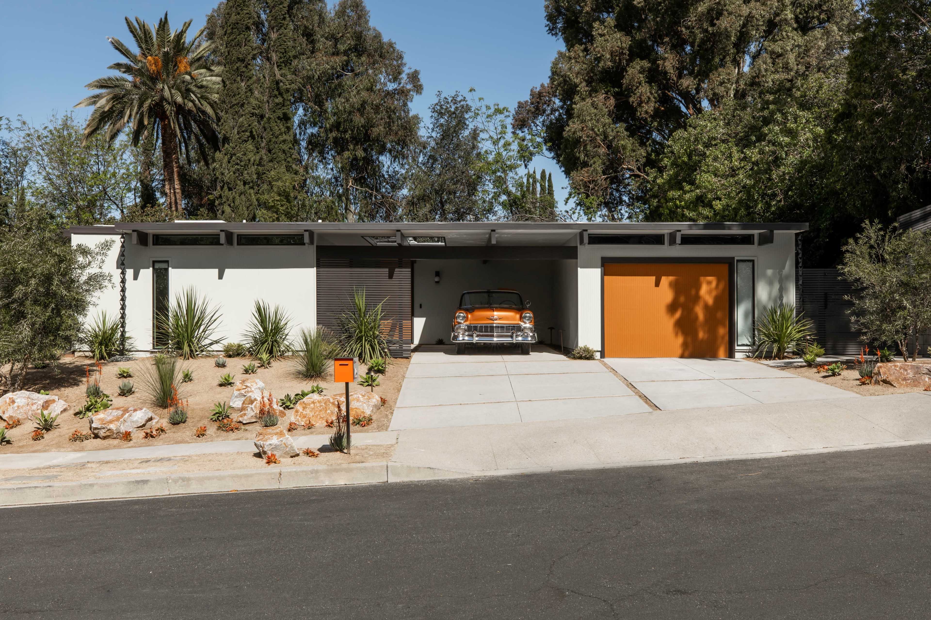 A modern house features a classic car parked in the driveway alongside a bright orange garage door and landscaped vegetation.
