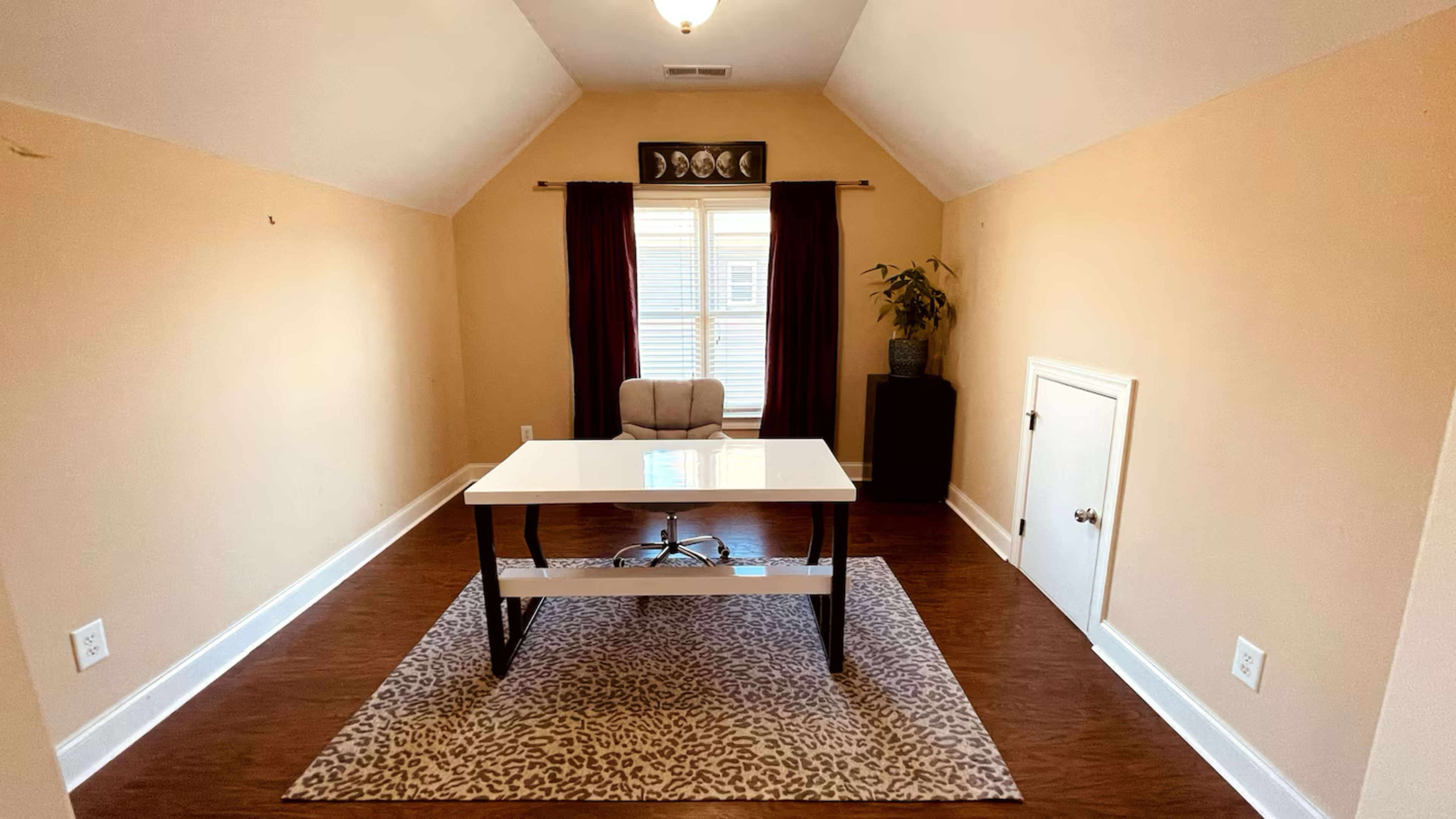 A spacious home office features a white desk and an ergonomic chair on a patterned rug, with a window dressed in dark curtains and a small plant in the corner.