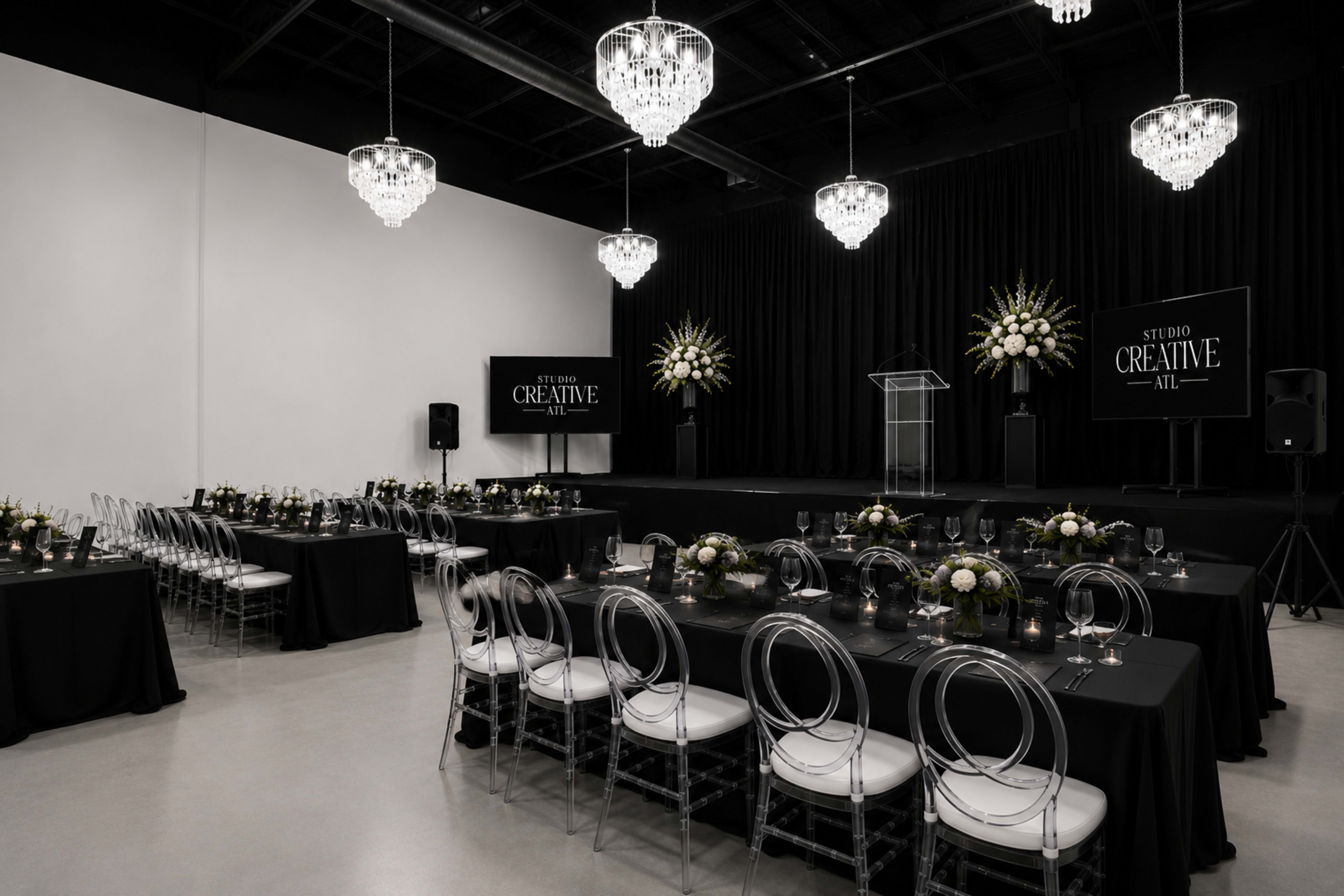 The image shows a modern event space set up for a gathering, featuring black tables with floral centerpieces, clear ghost chairs, and chandeliers hanging from the ceiling.