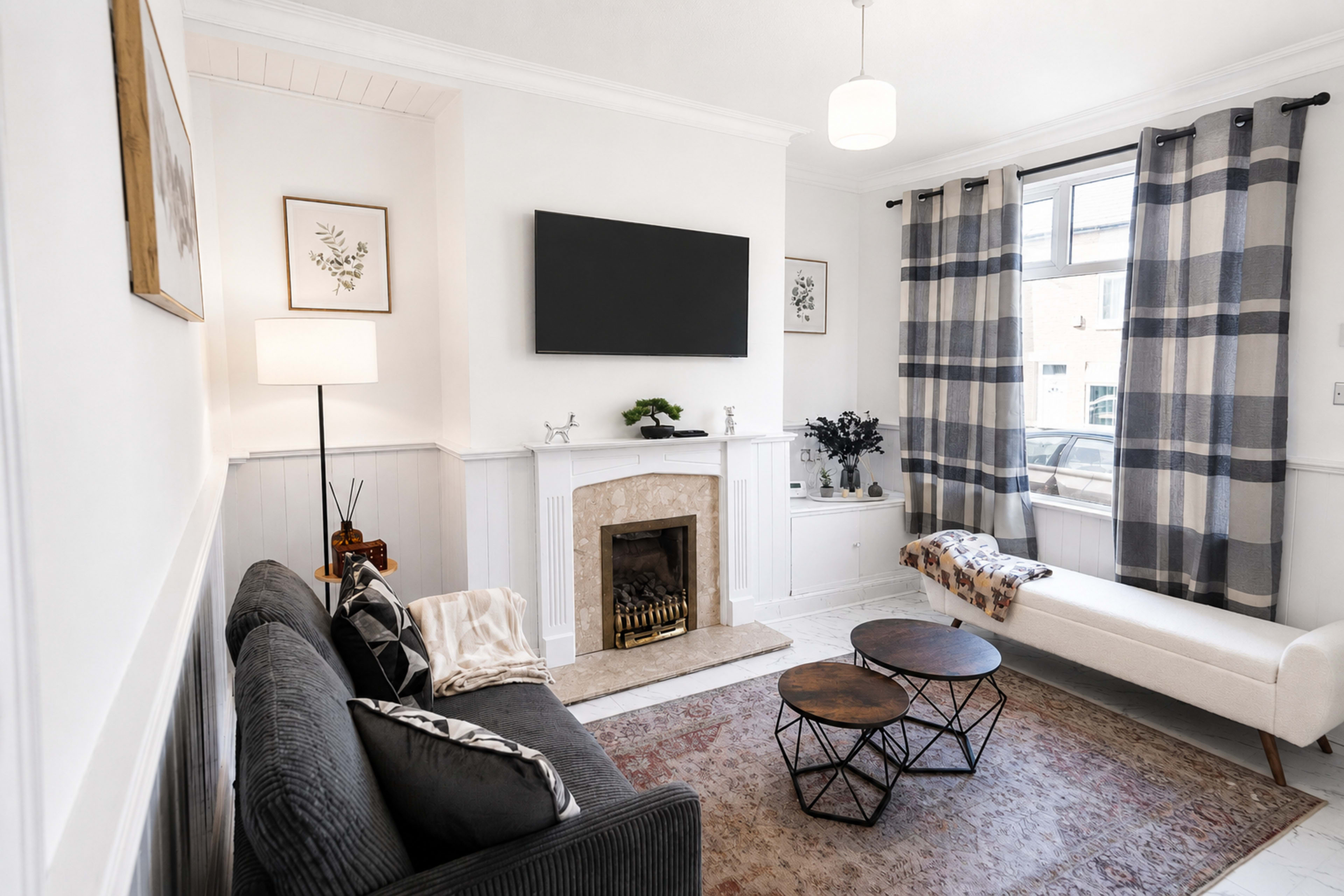 The image shows a modern living room featuring a grey sofa, a white loveseat, a decorative fireplace, two coffee tables, and large windows with plaid curtains.