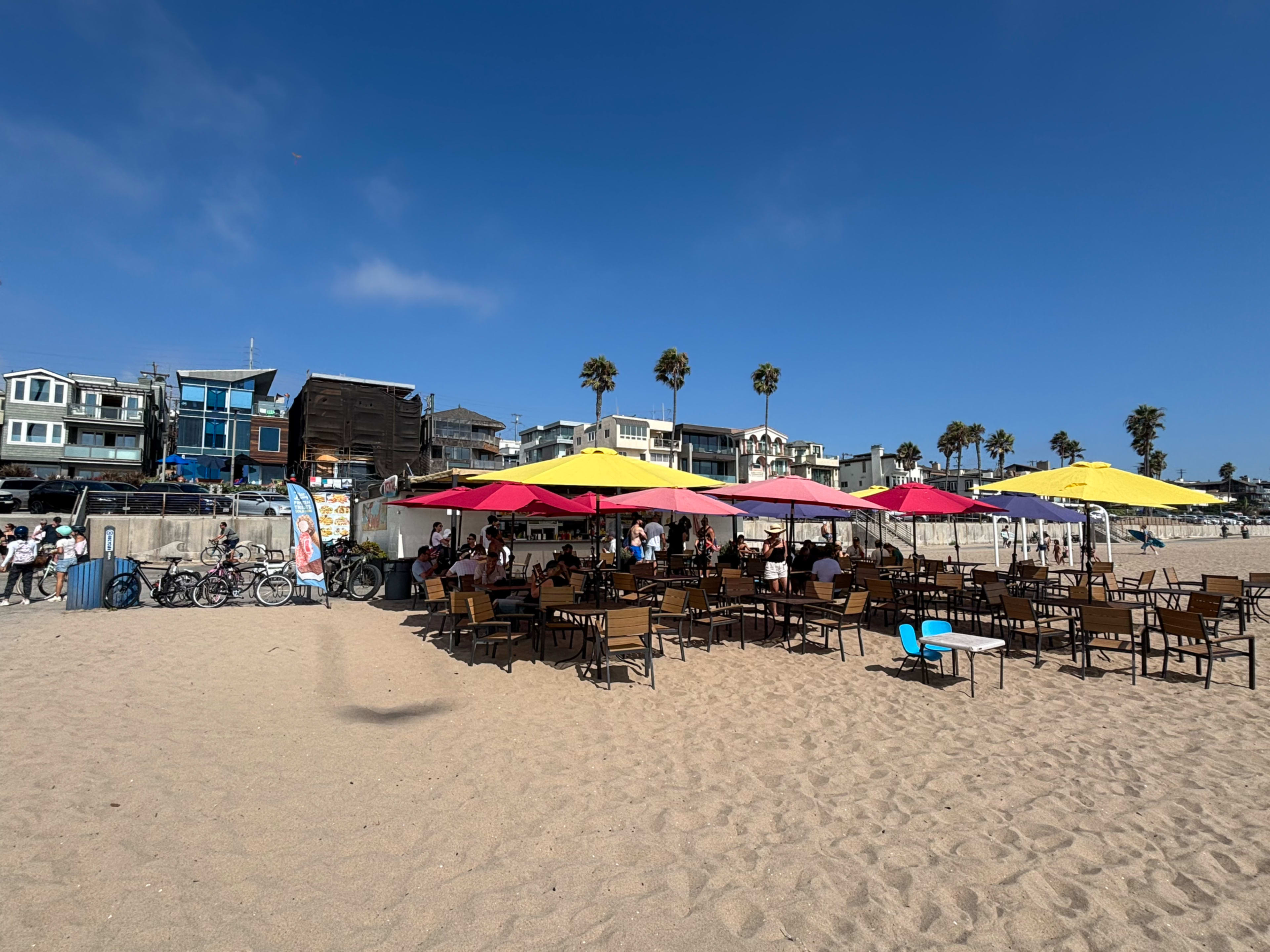 A beachside cafe with outdoor seating and colorful umbrellas is bustling with patrons while palm trees and beach houses line the background.