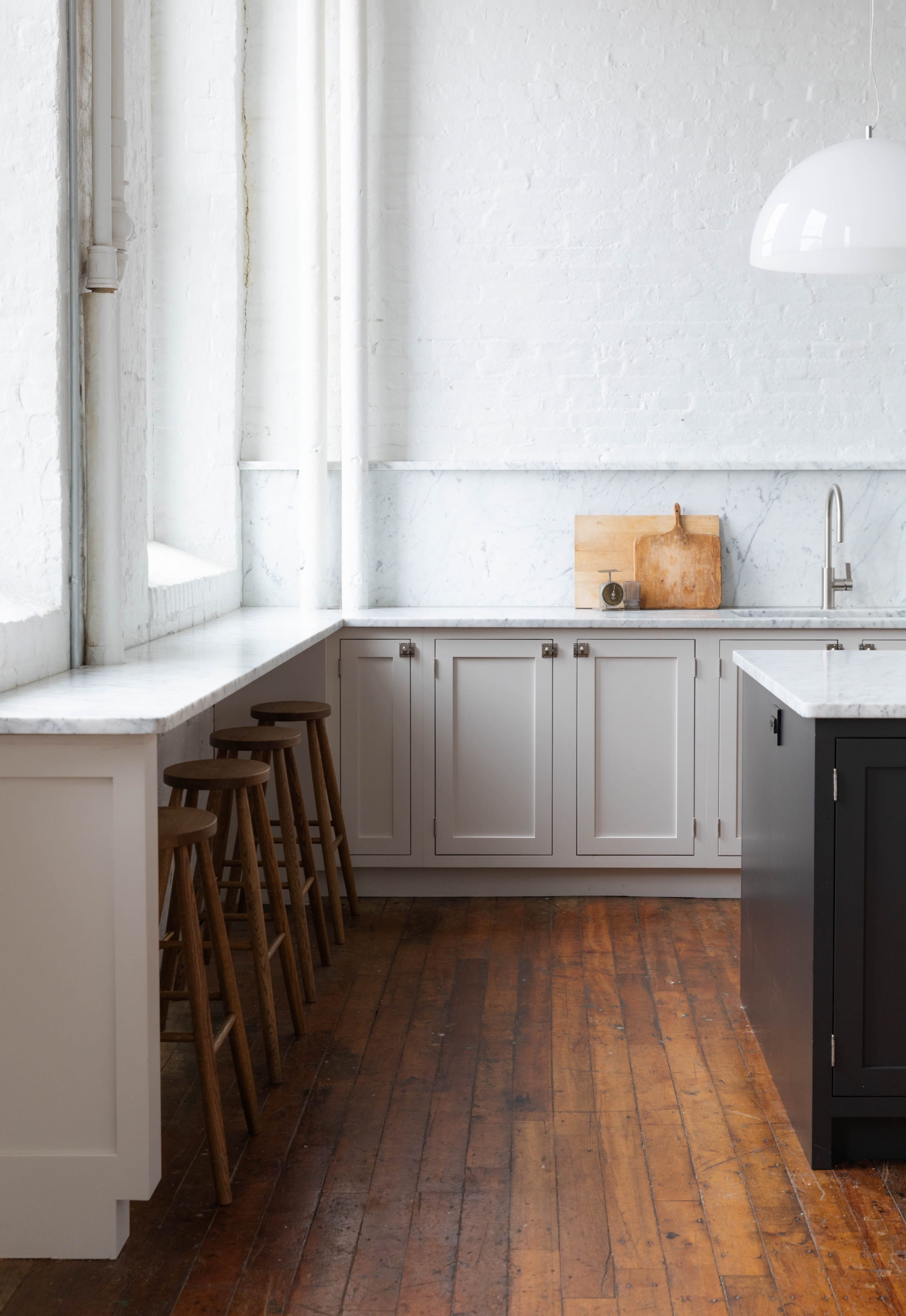 A modern kitchen features light-colored cabinetry, a marble countertop, wooden floors, and three tall stools along one side.