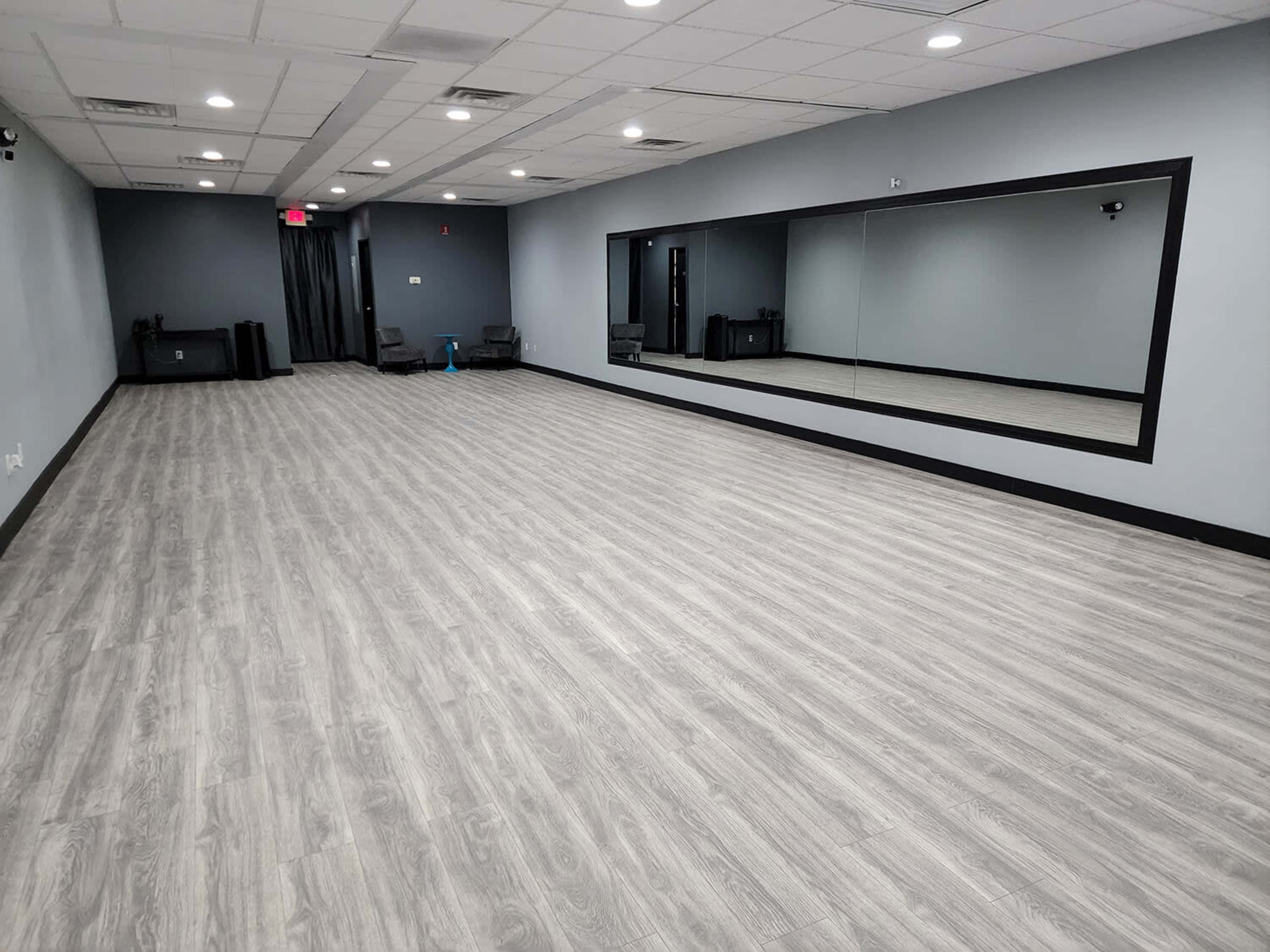 A spacious, empty room with gray walls, a wooden floor, and a large mirror along one side.