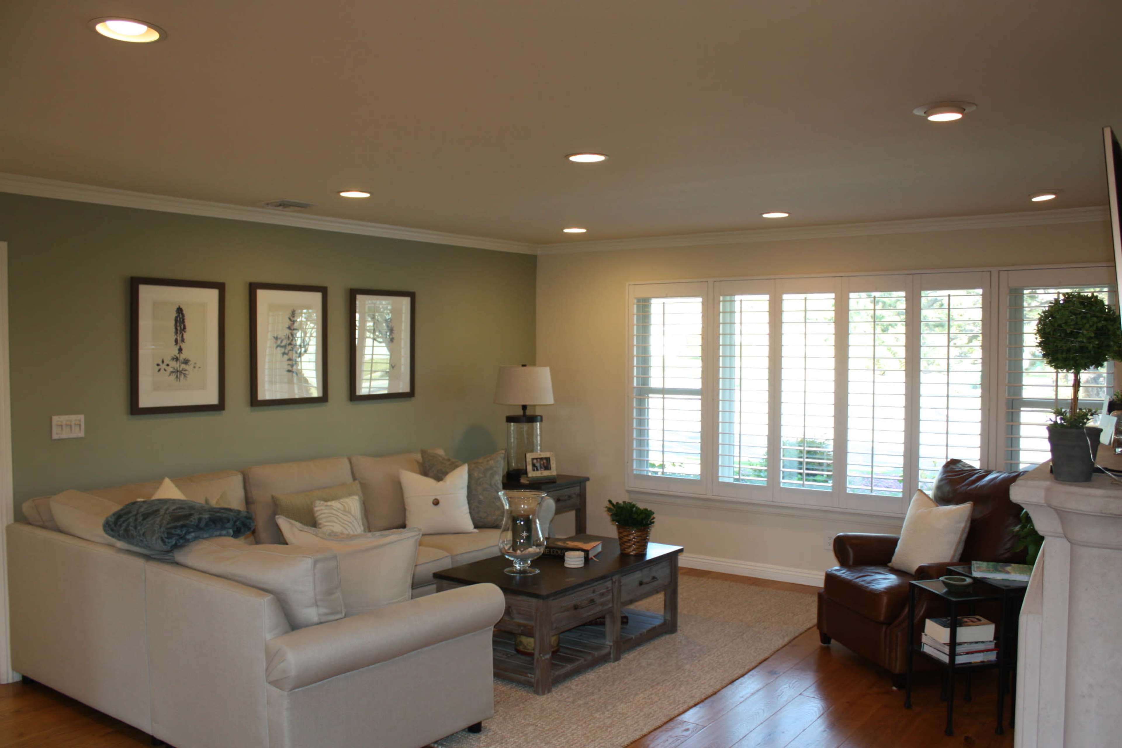 A light-filled living room features a beige sectional sofa, a wooden coffee table, and a chair beside a window with plantation shutters.