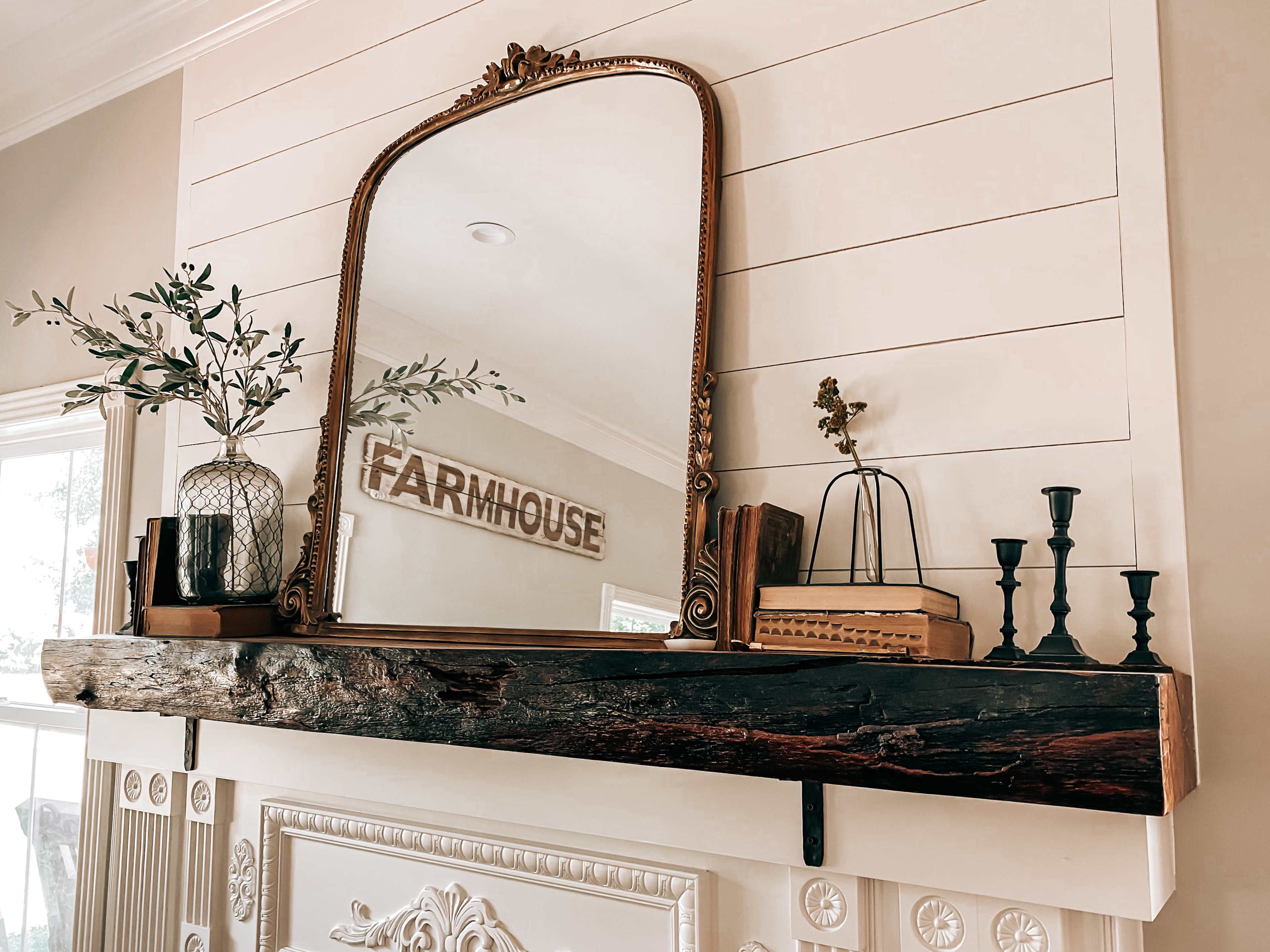 A wooden mantel with a vintage mirror, decorative plants, and various accents is mounted on a white paneled wall.