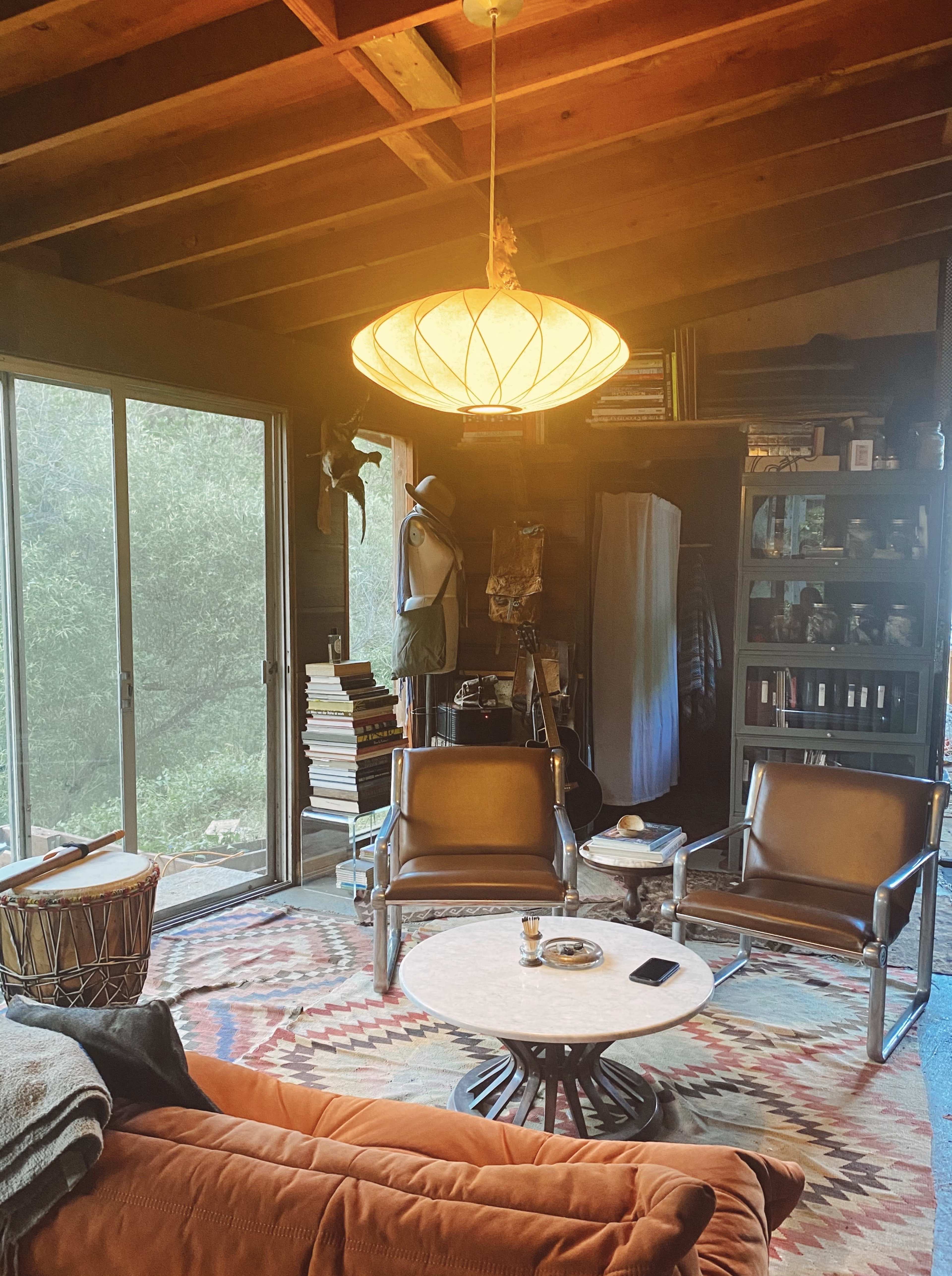 A cozy living room with a large window, a circular coffee table, two leather chairs, a sofa, and shelves filled with books and various items.