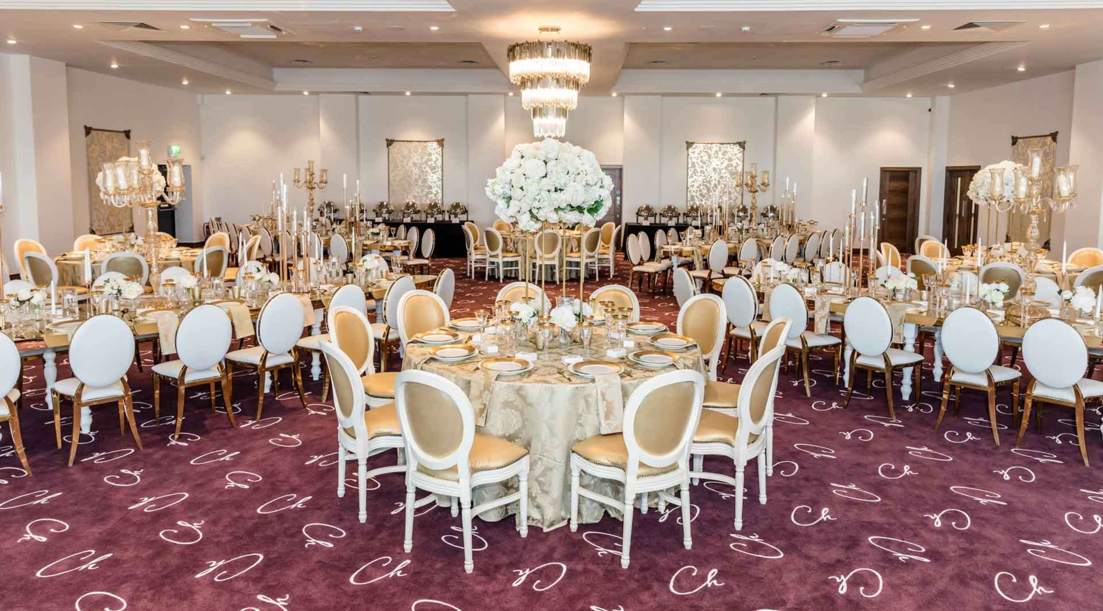 An elegantly set banquet hall featuring round tables with floral centerpieces, surrounded by white chairs on a dark carpet.