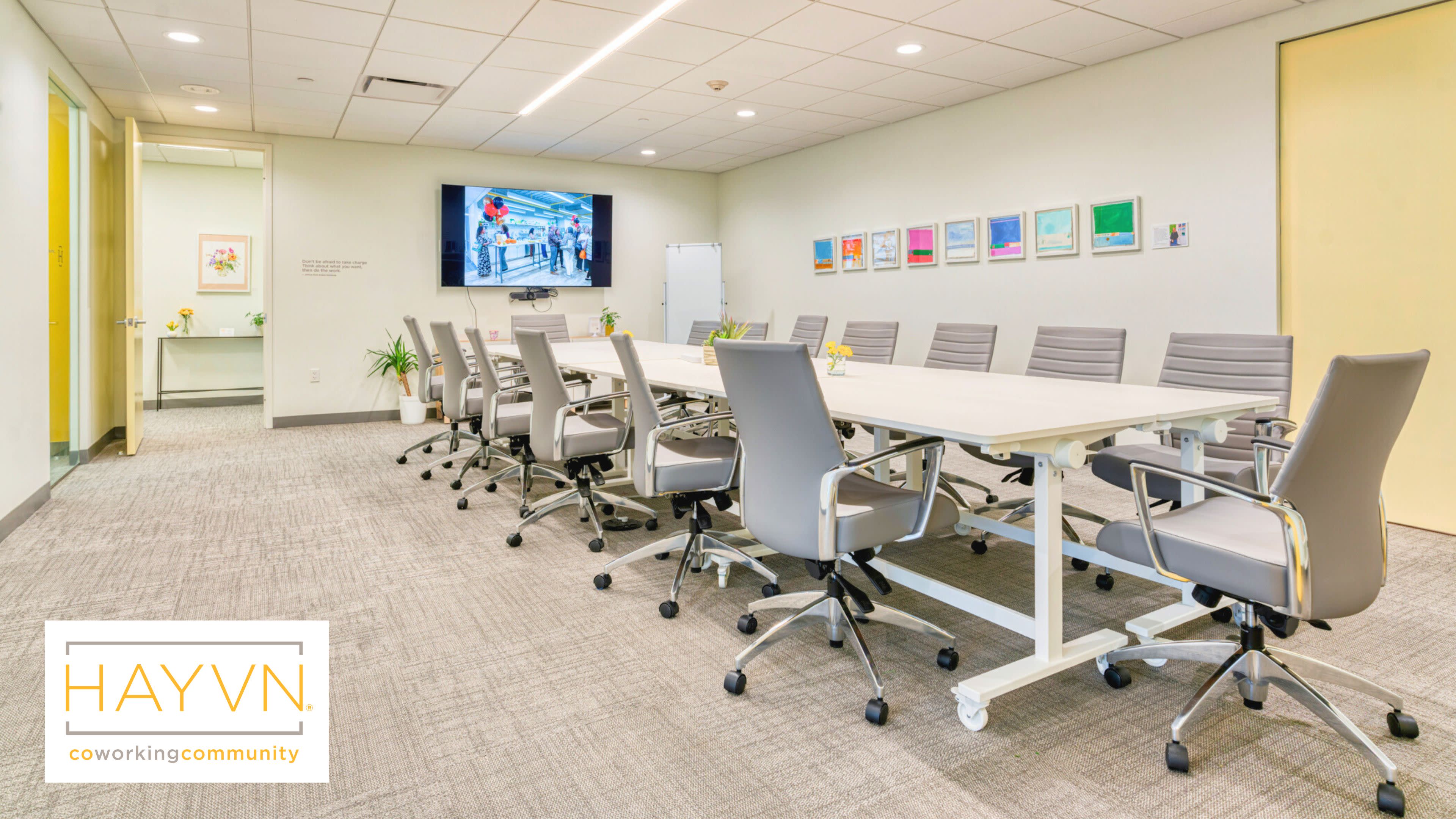 The image shows a modern conference room with a long table and ergonomic chairs, featuring framed artwork on the walls and a large screen at one end.