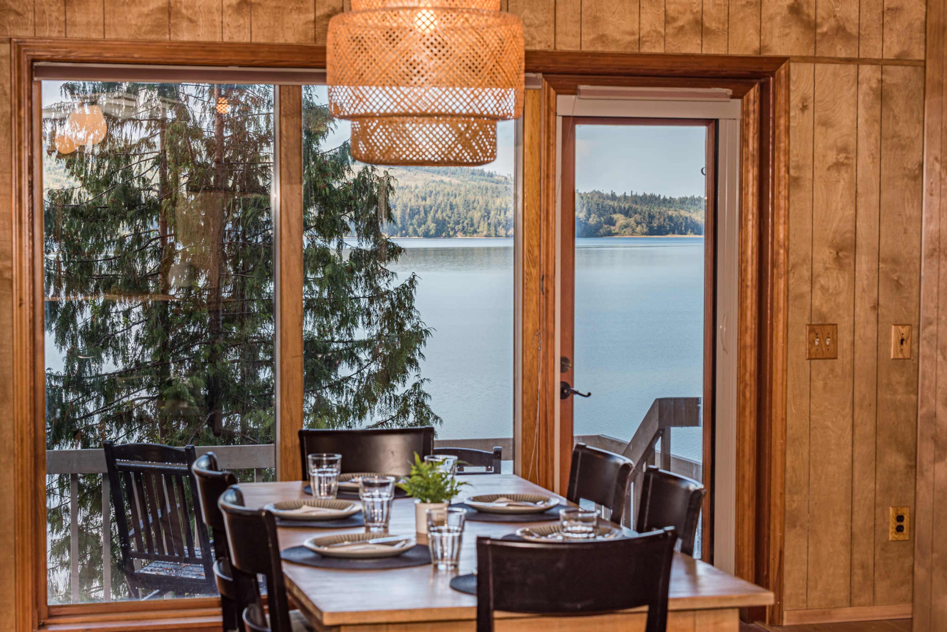 A dining table set with plates and glasses overlooks a serene lake through large windows framed by wooden walls.