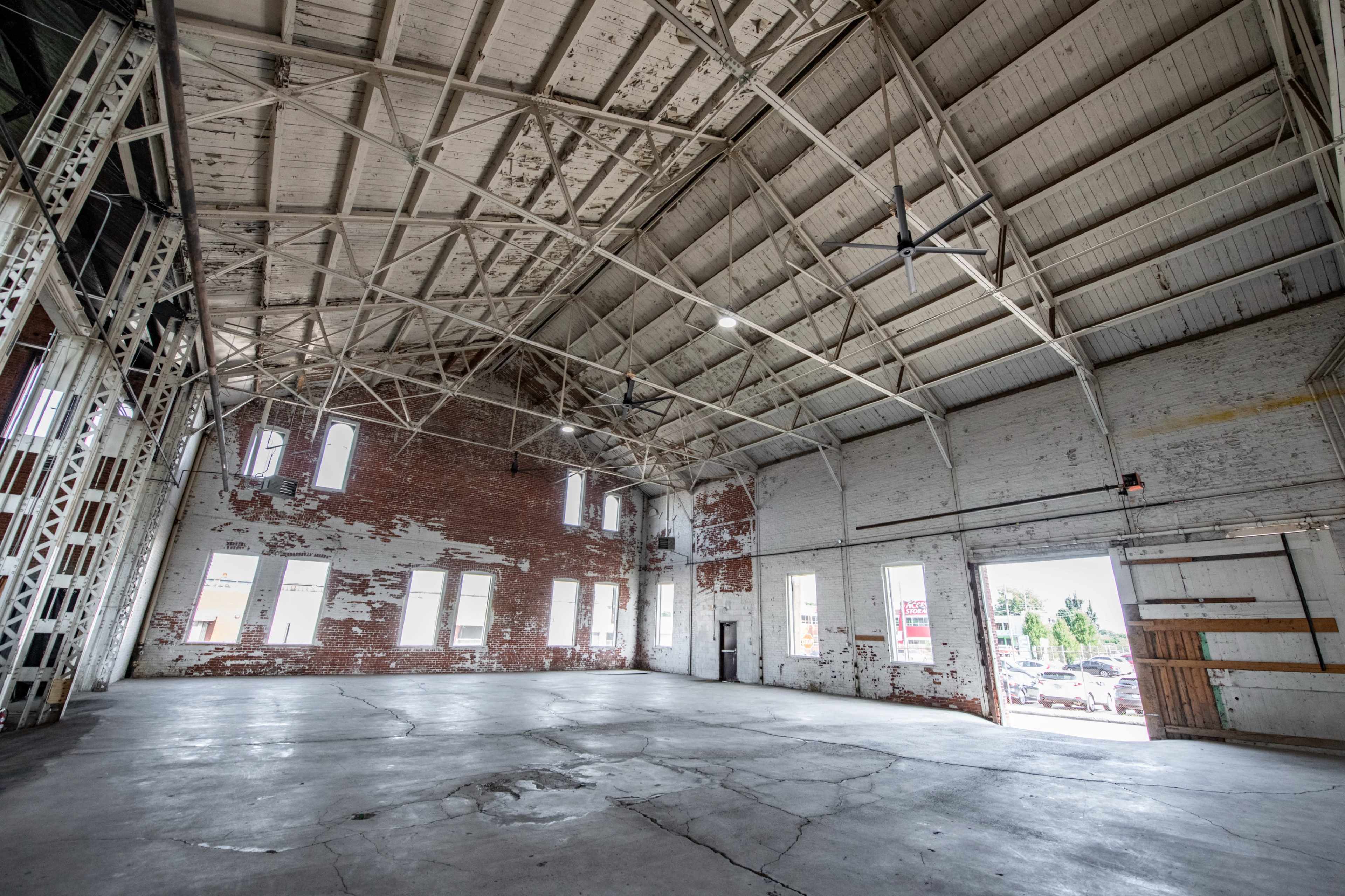 The image shows a large, empty warehouse with a high, metal-truss ceiling and exposed brick walls.