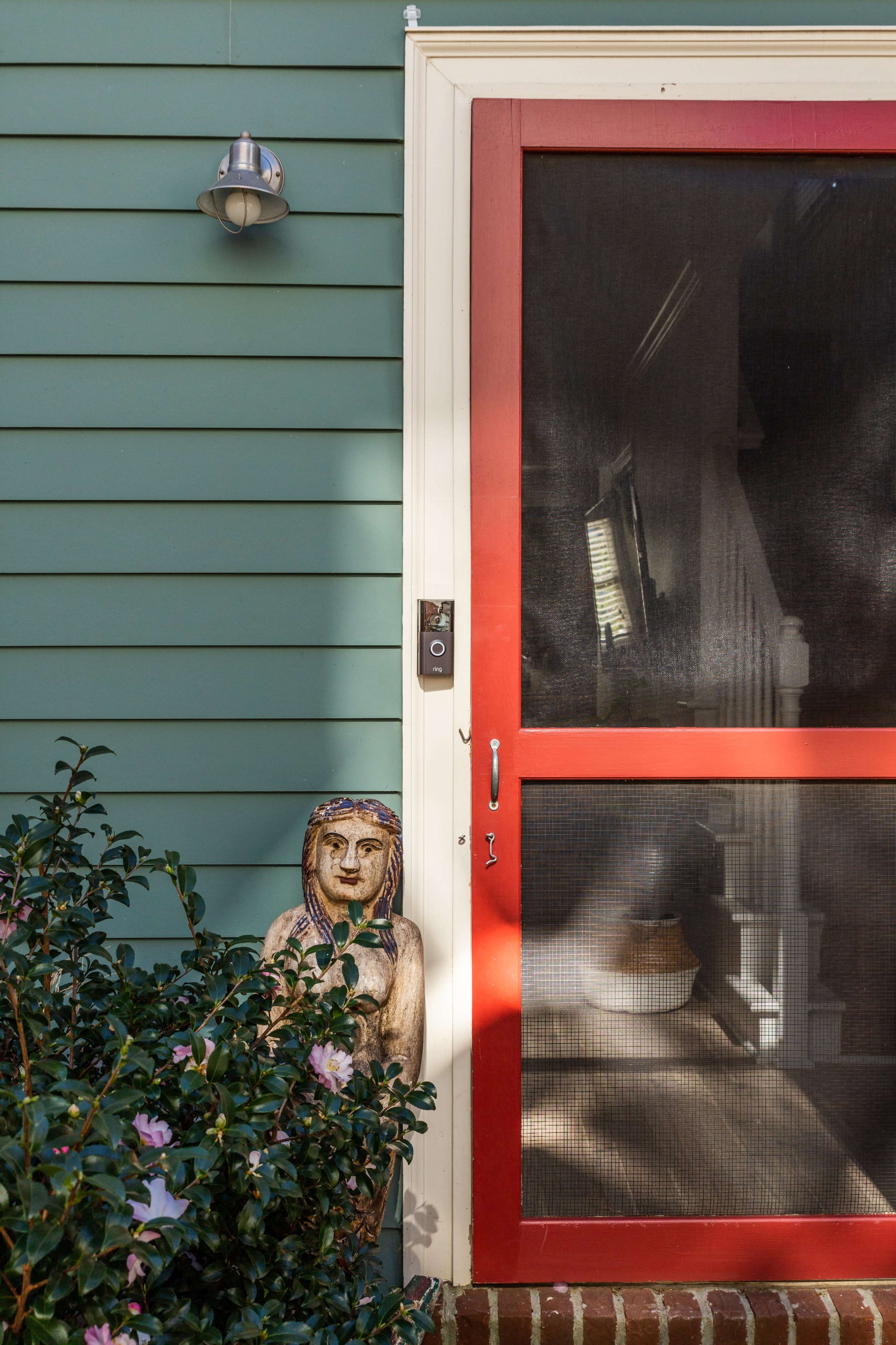 A carved figure is positioned beside a red screen door, framed by green siding and a light fixture above.