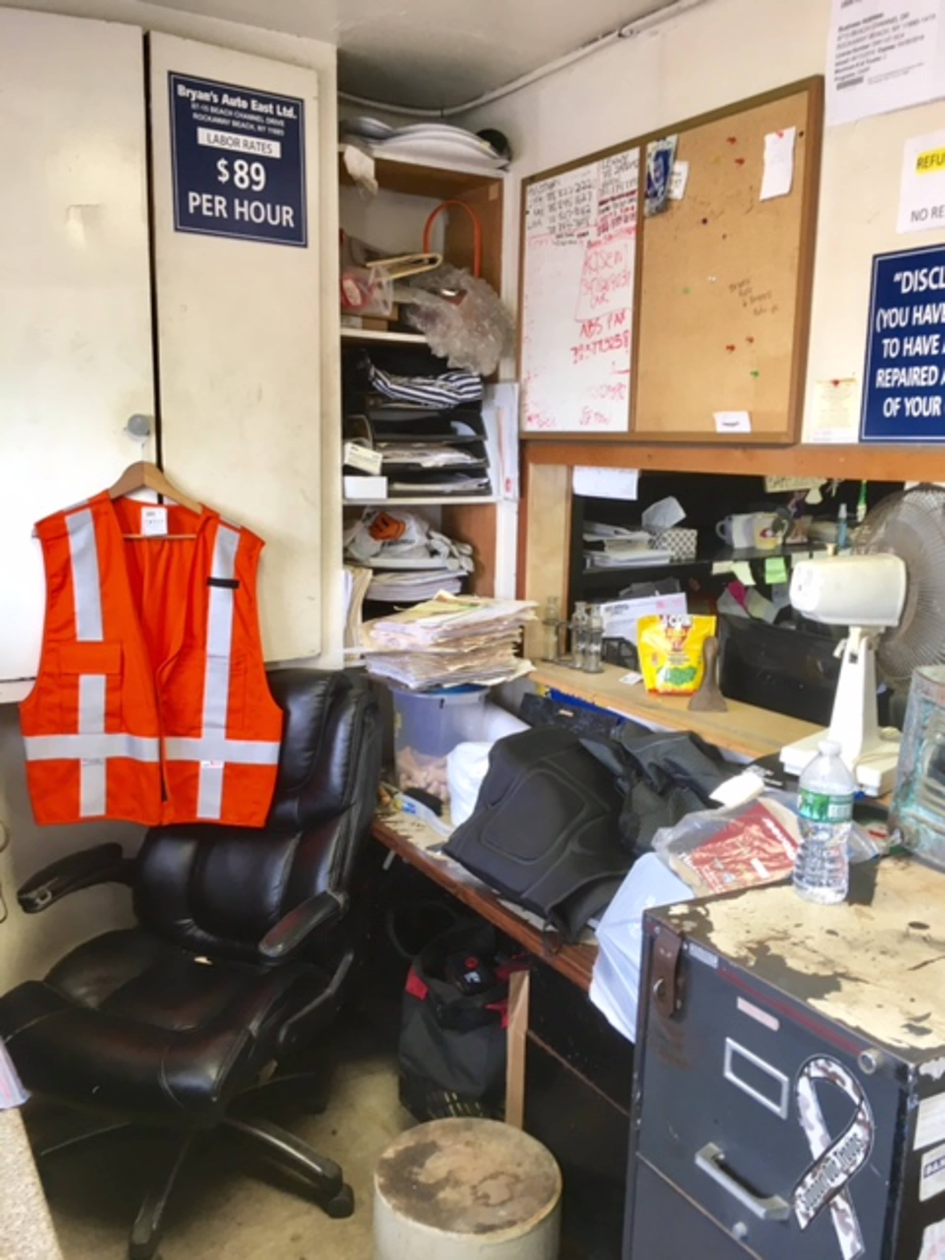 A cluttered office space with a chair, an orange safety vest hanging, and various papers and items scattered across a desk.