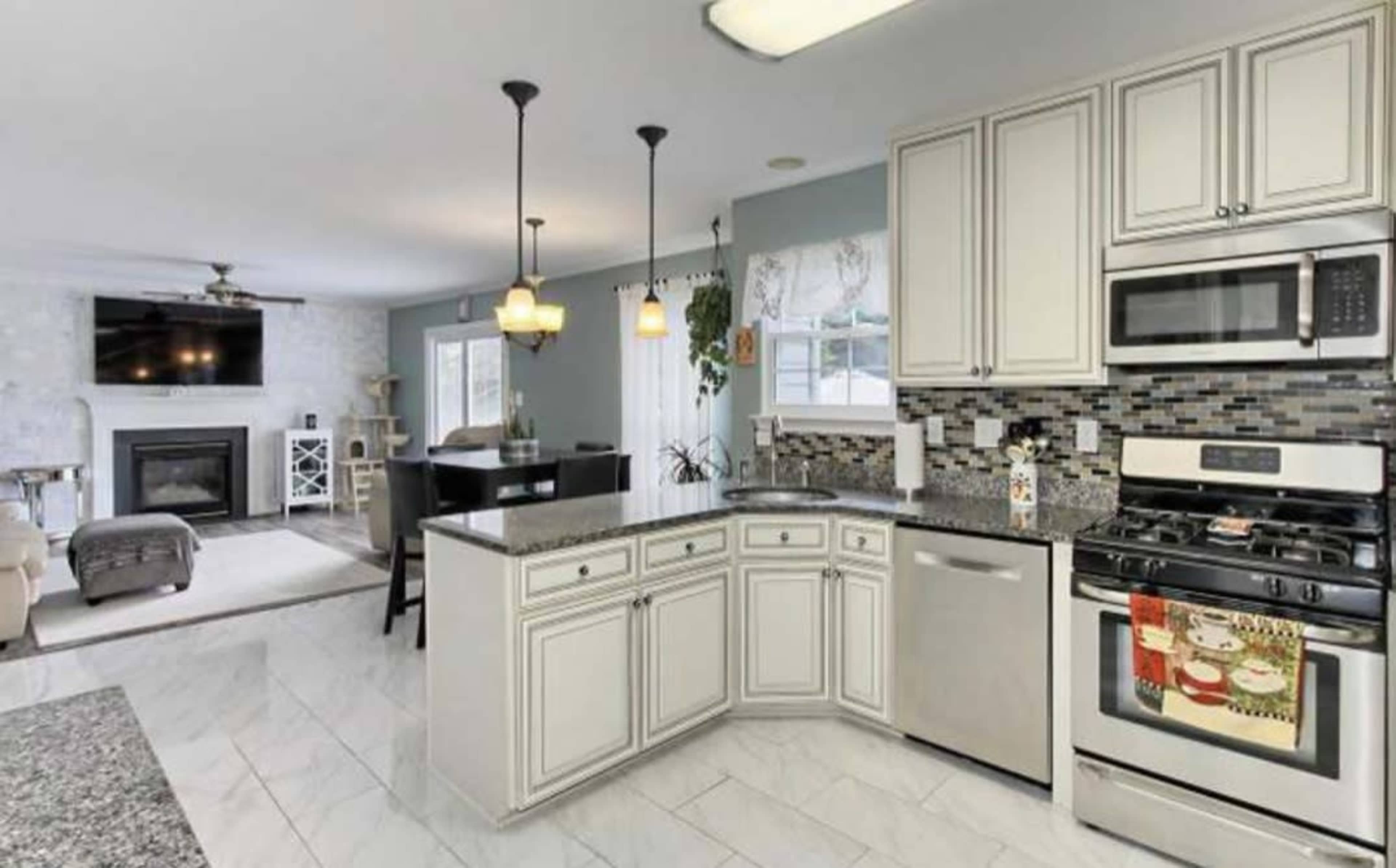 A modern kitchen features white cabinets, a granite countertop, stainless steel appliances, and a dining area with a view of a living room and fireplace.
