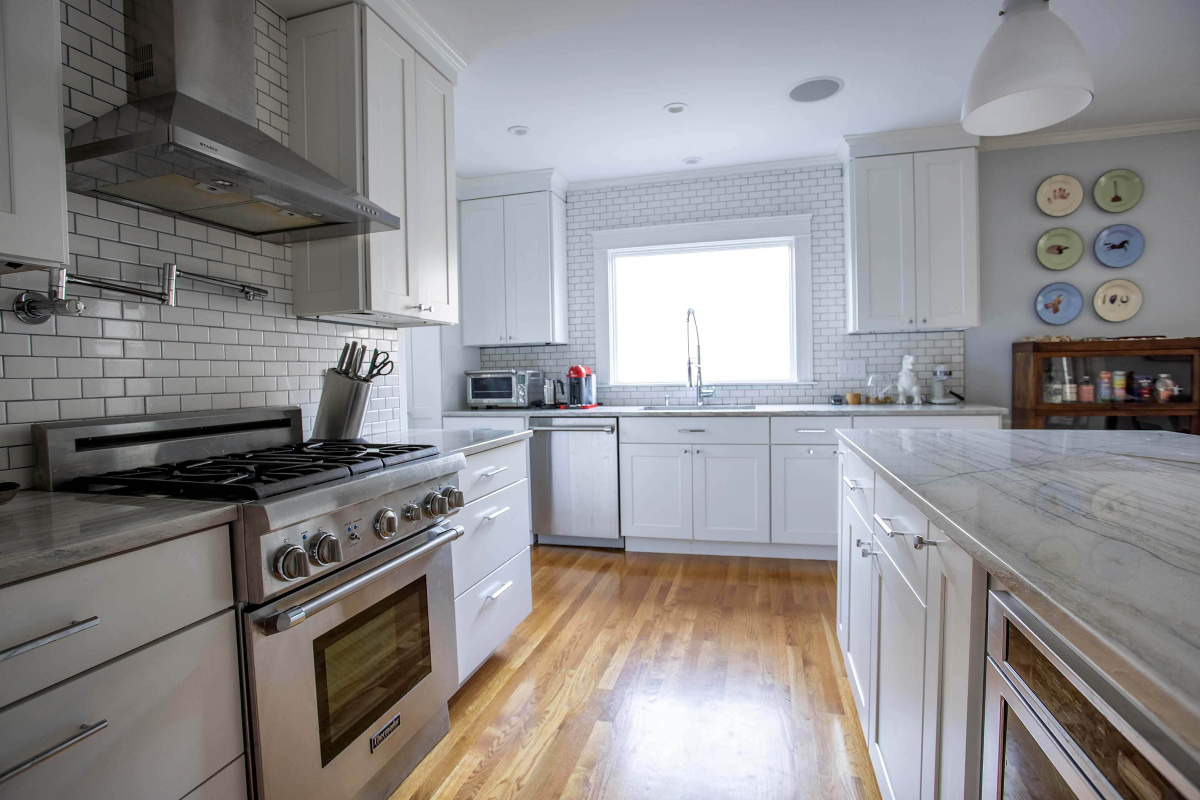 A bright kitchen features white cabinets, a stainless steel oven, and a large window, with marble countertops and wooden flooring.