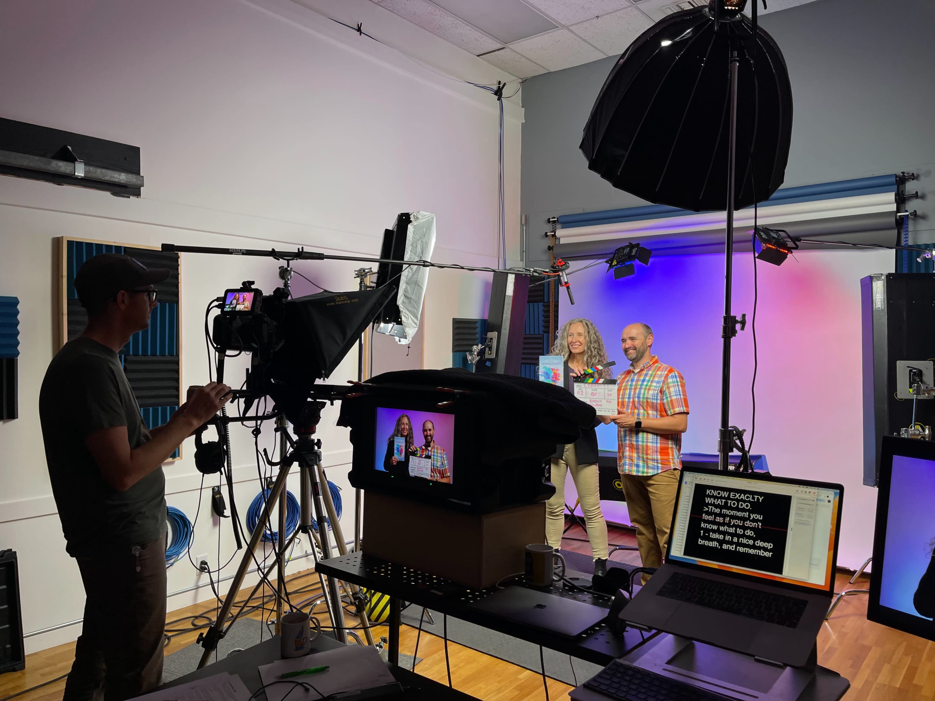 A video production crew sets up equipment while two people hold awards in front of a colorful backdrop.