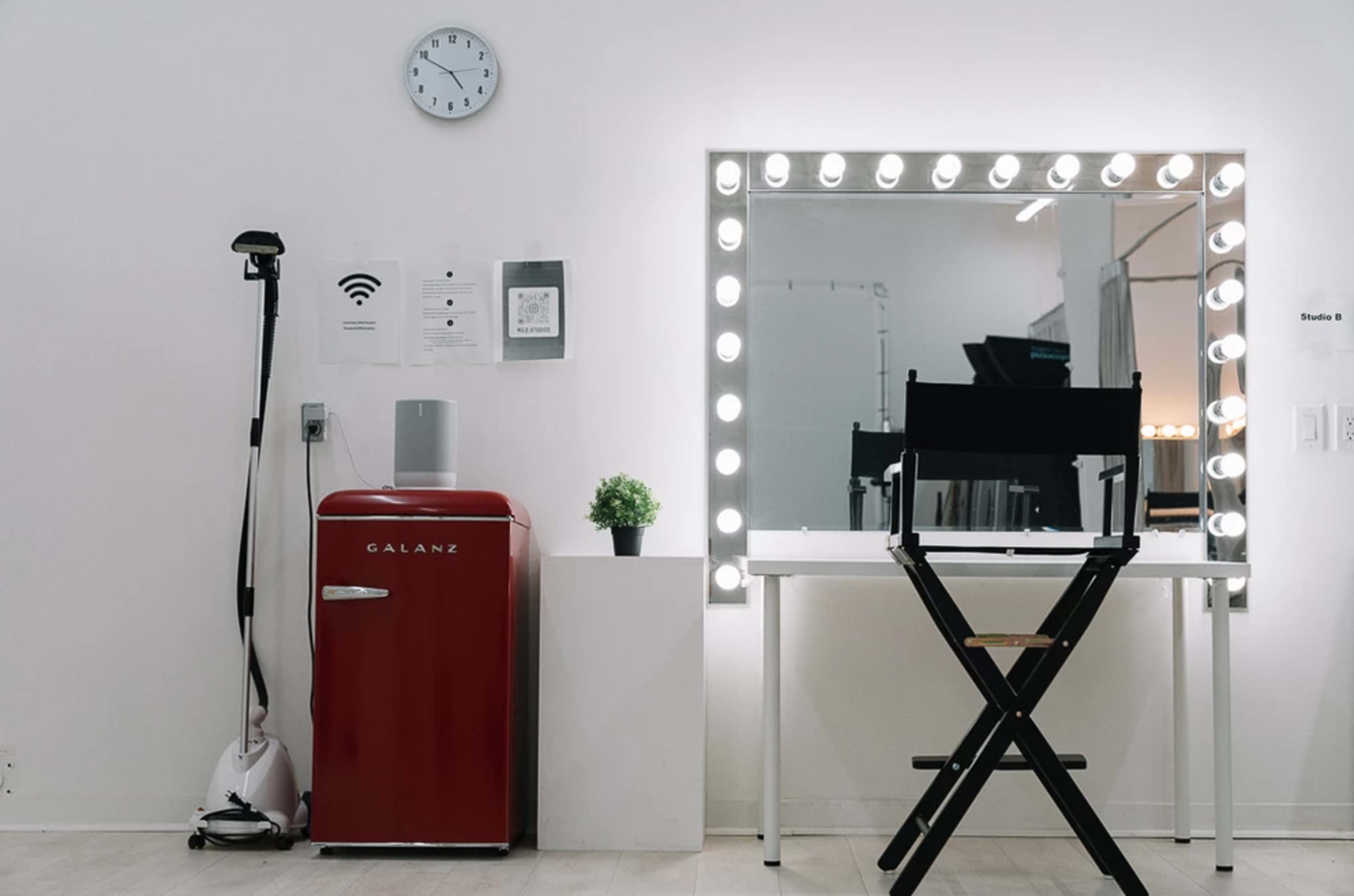 The scene features a modern dressing area with a red refrigerator, a tall mirror surrounded by lights, a director's chair, and a clock on the wall.