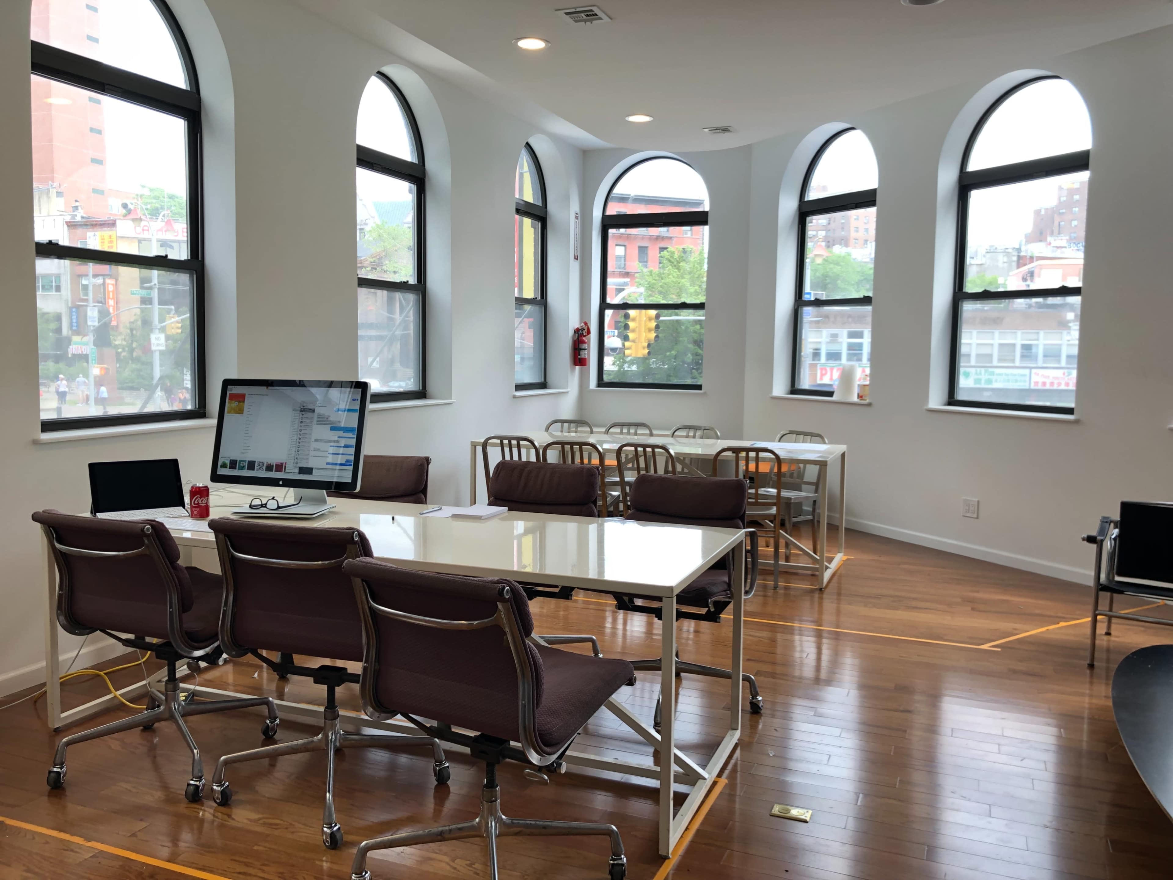 The image shows a bright, modern workspace with several tables and chairs arranged near large, arched windows.