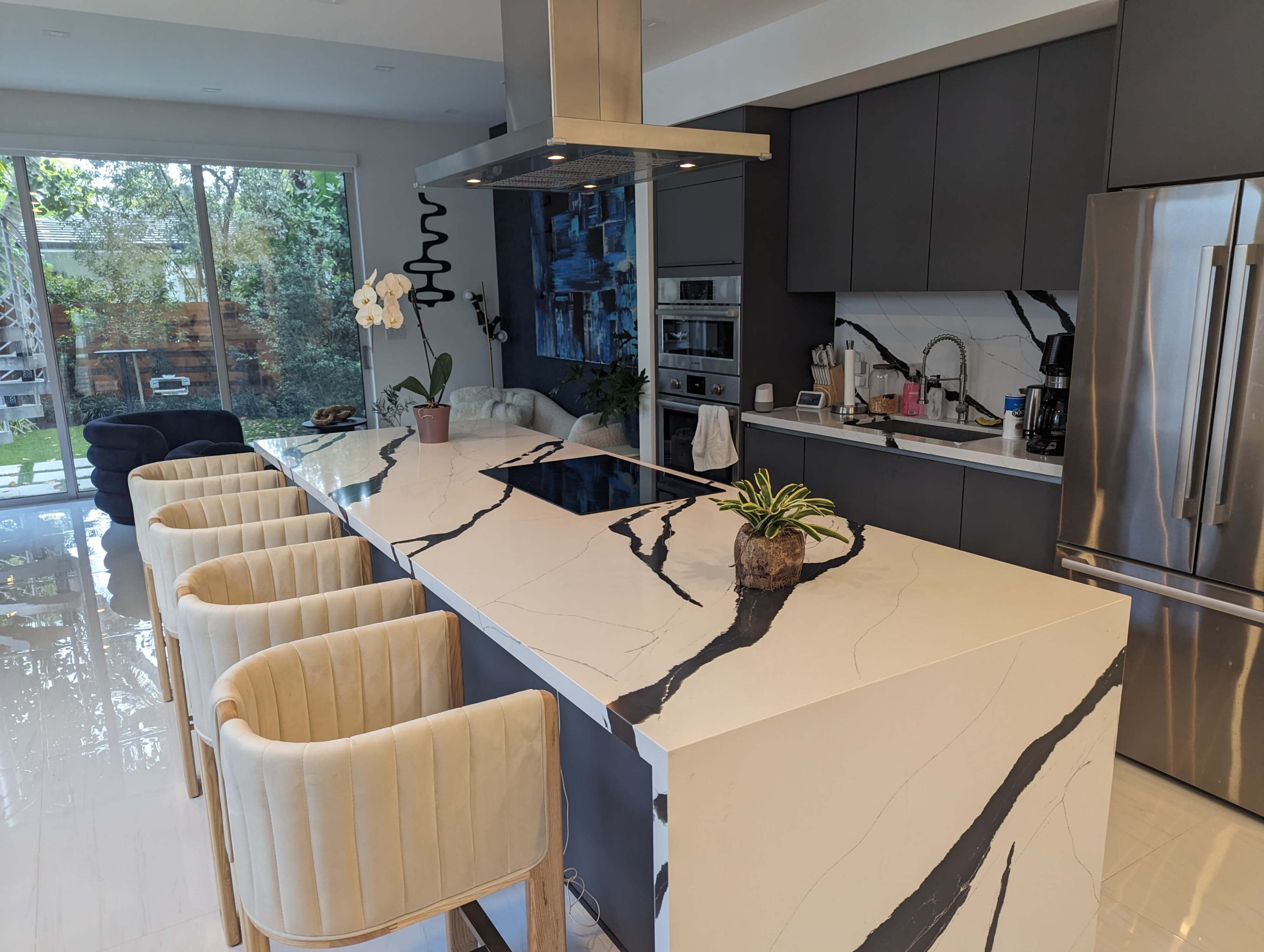 A contemporary kitchen features a large marble island with four cream-colored bar stools, modern appliances, and a view of a landscaped backyard through large windows.