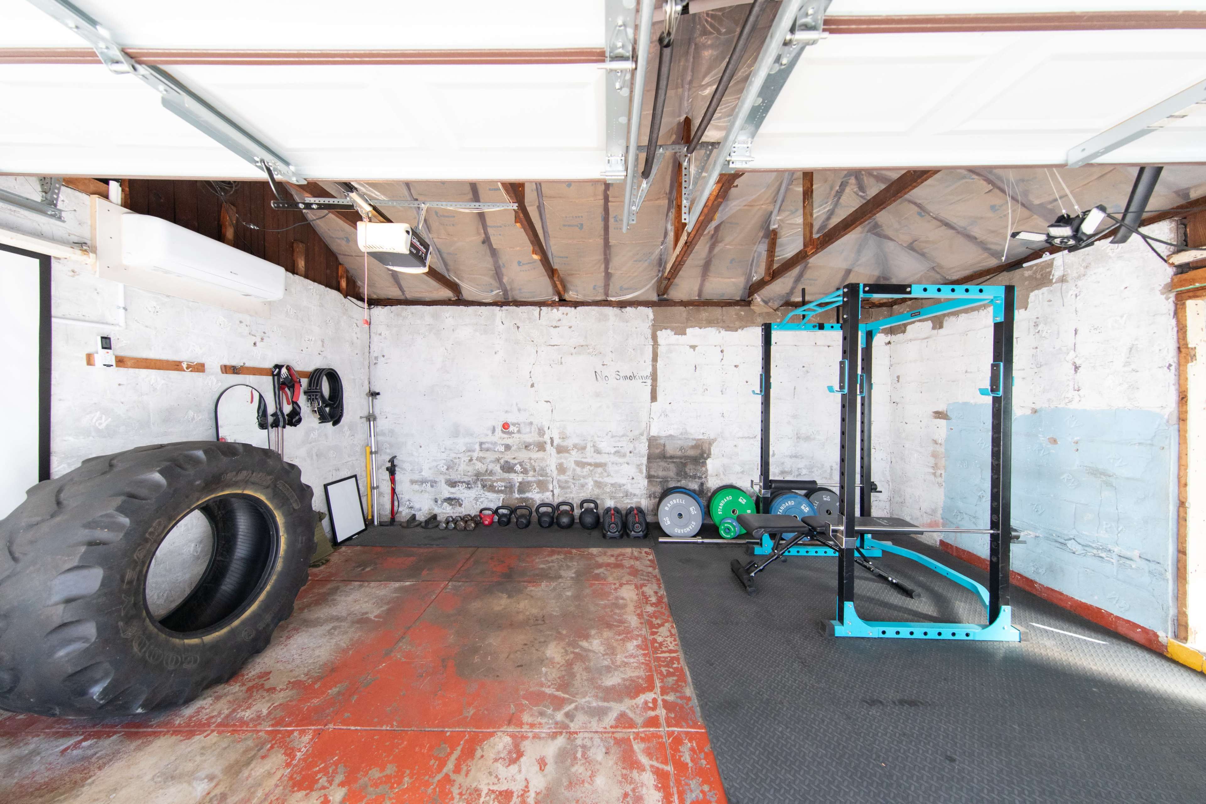 The image shows an empty garage space with a tire, dumbbells, and a weightlifting rack against a concrete wall.