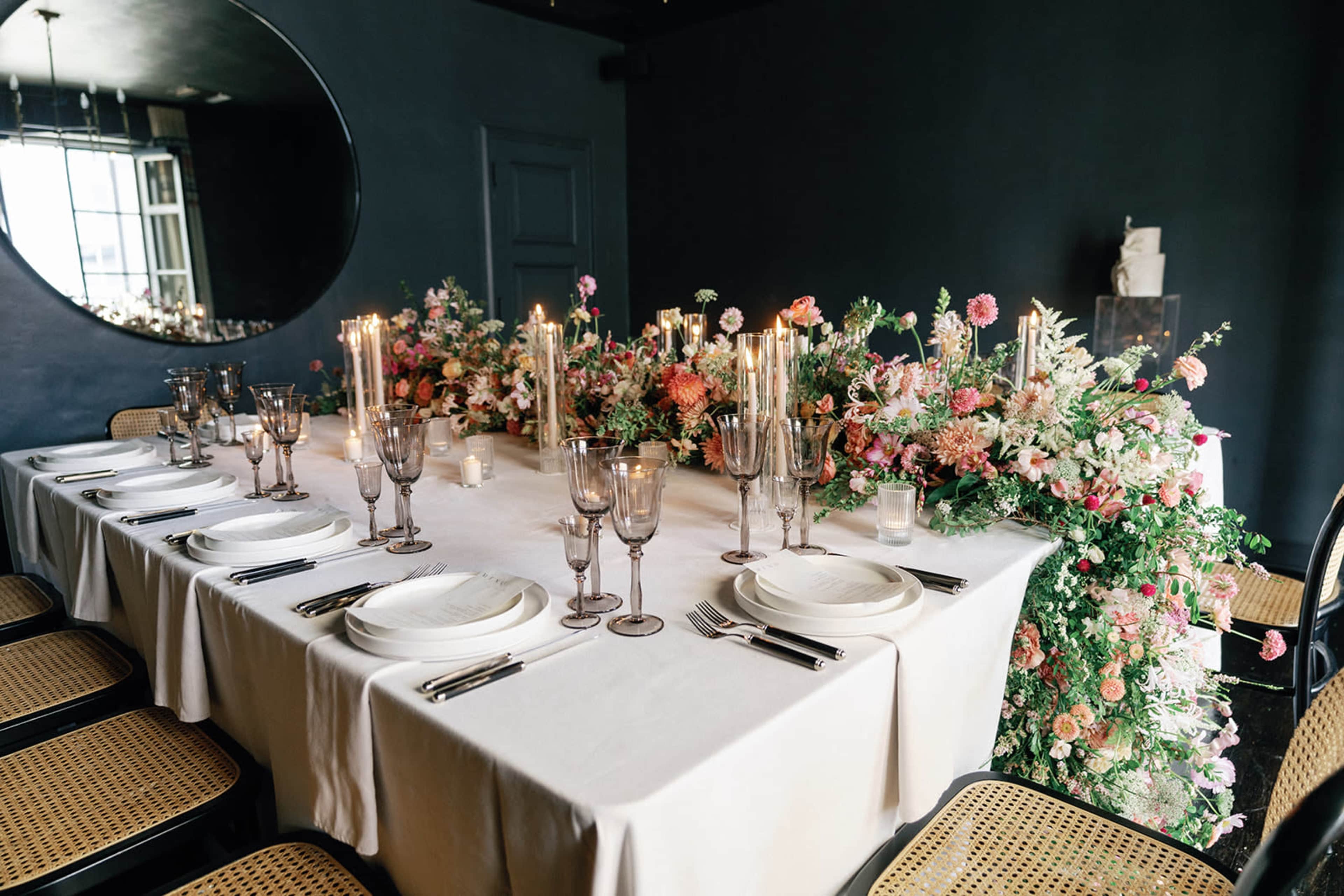 A long, elegantly set dining table is adorned with flowers and candles, located in a dark-colored room with a round mirror reflecting the decor.