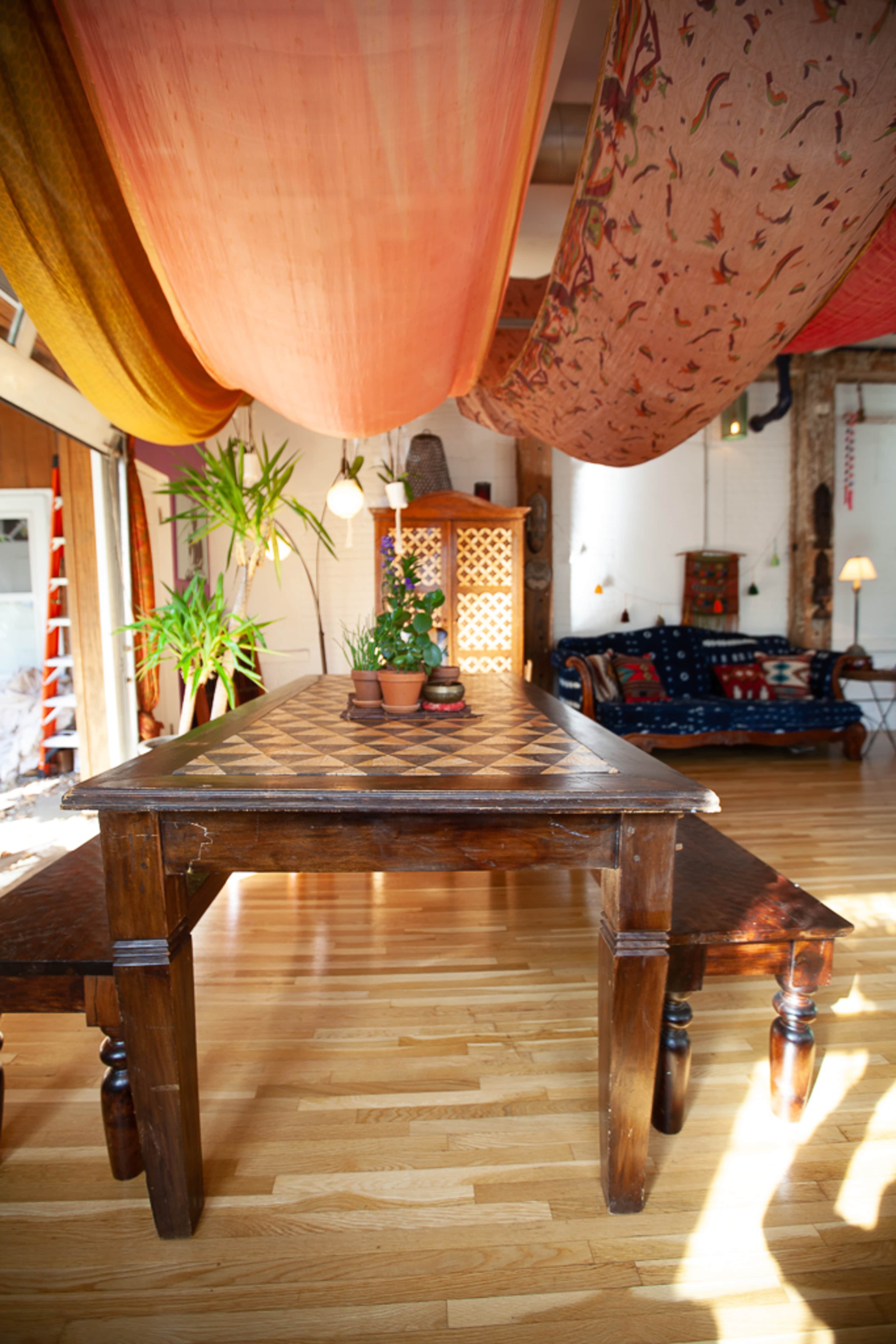 A wooden table sits in a brightly lit room adorned with colorful drapes and potted plants.