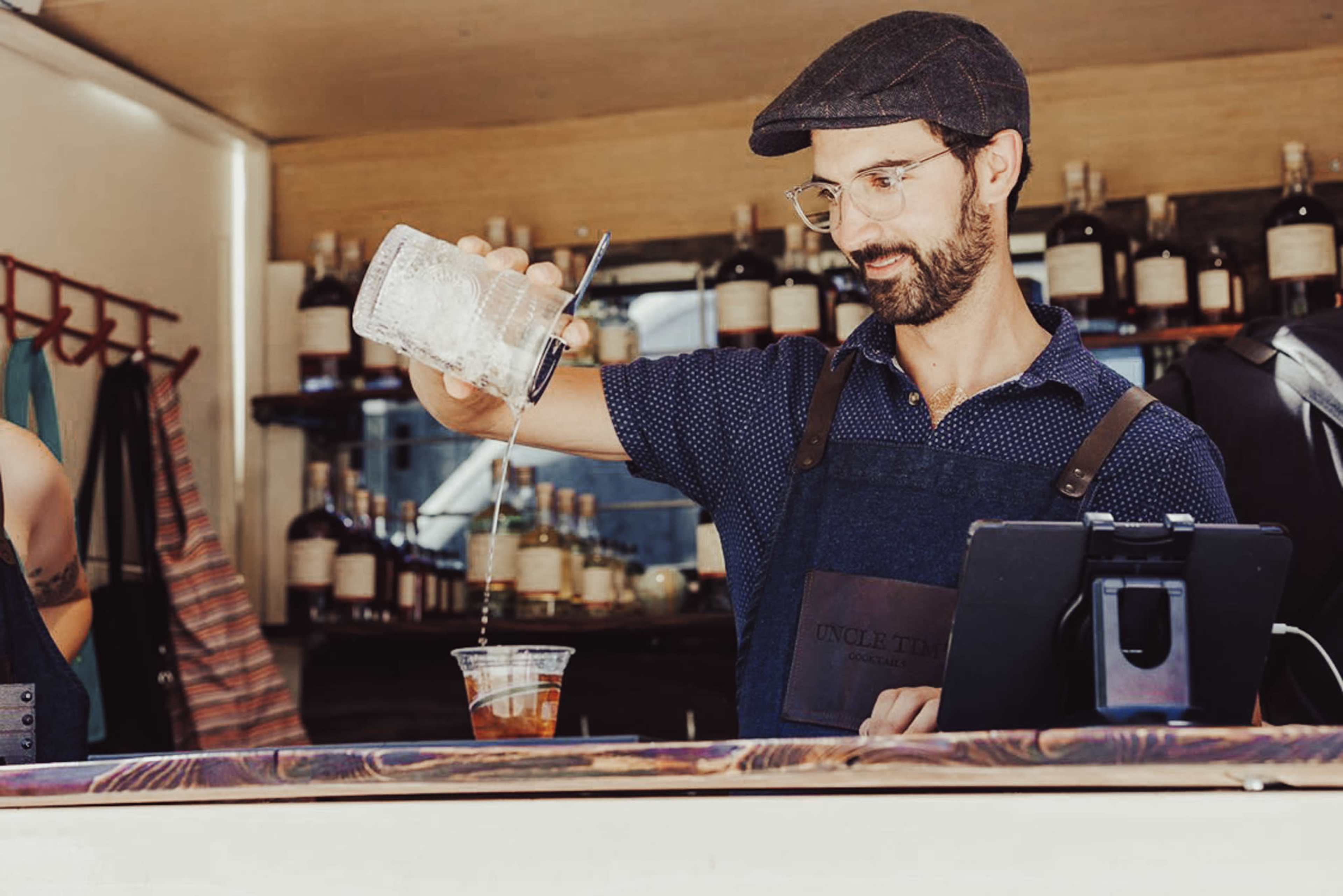 A bartender in a flat cap pours a drink into a plastic cup at a bar surrounded by bottles.