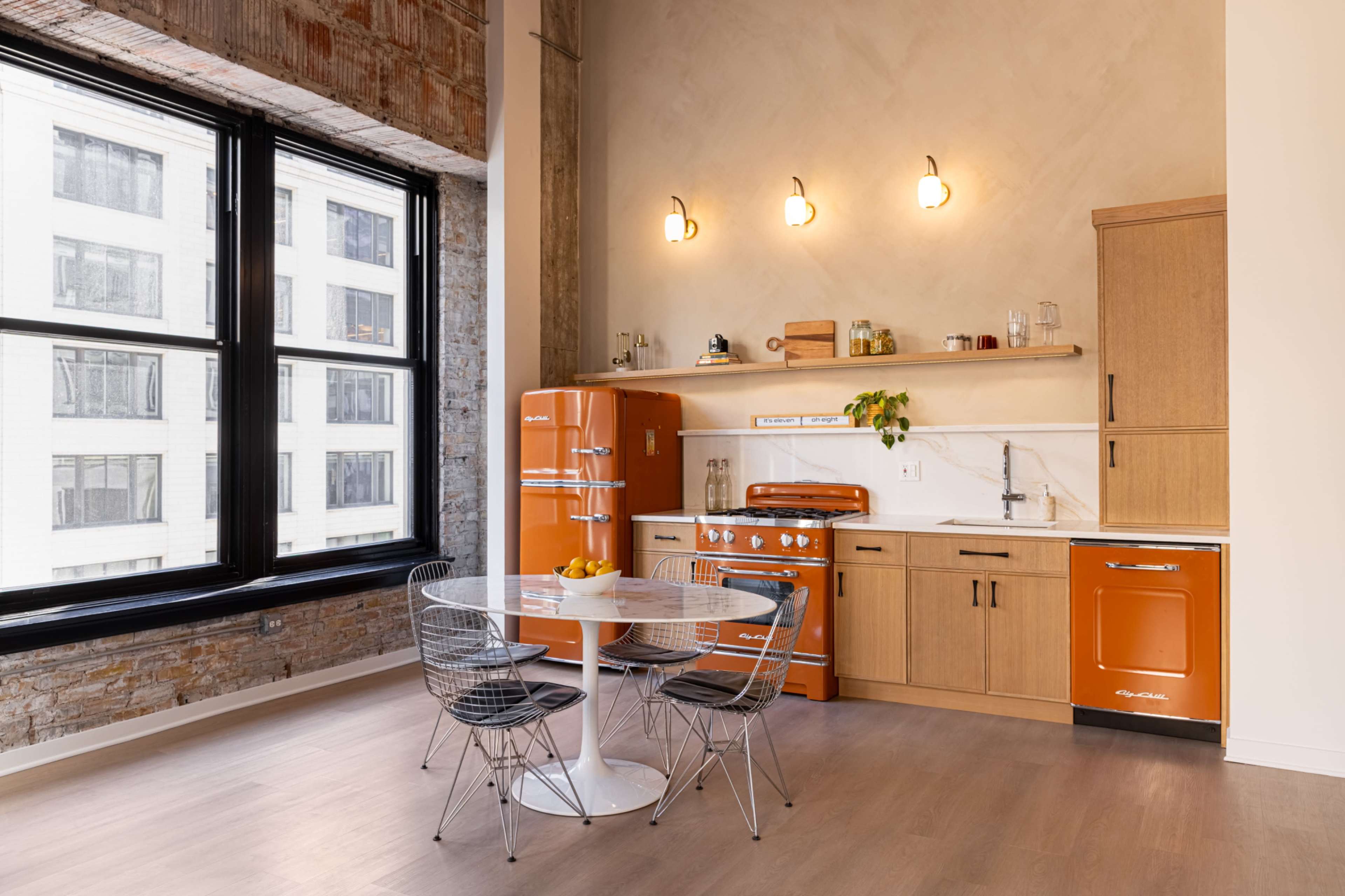 The image shows a modern kitchen with orange appliances, a round dining table, and large windows that allow natural light to enter.