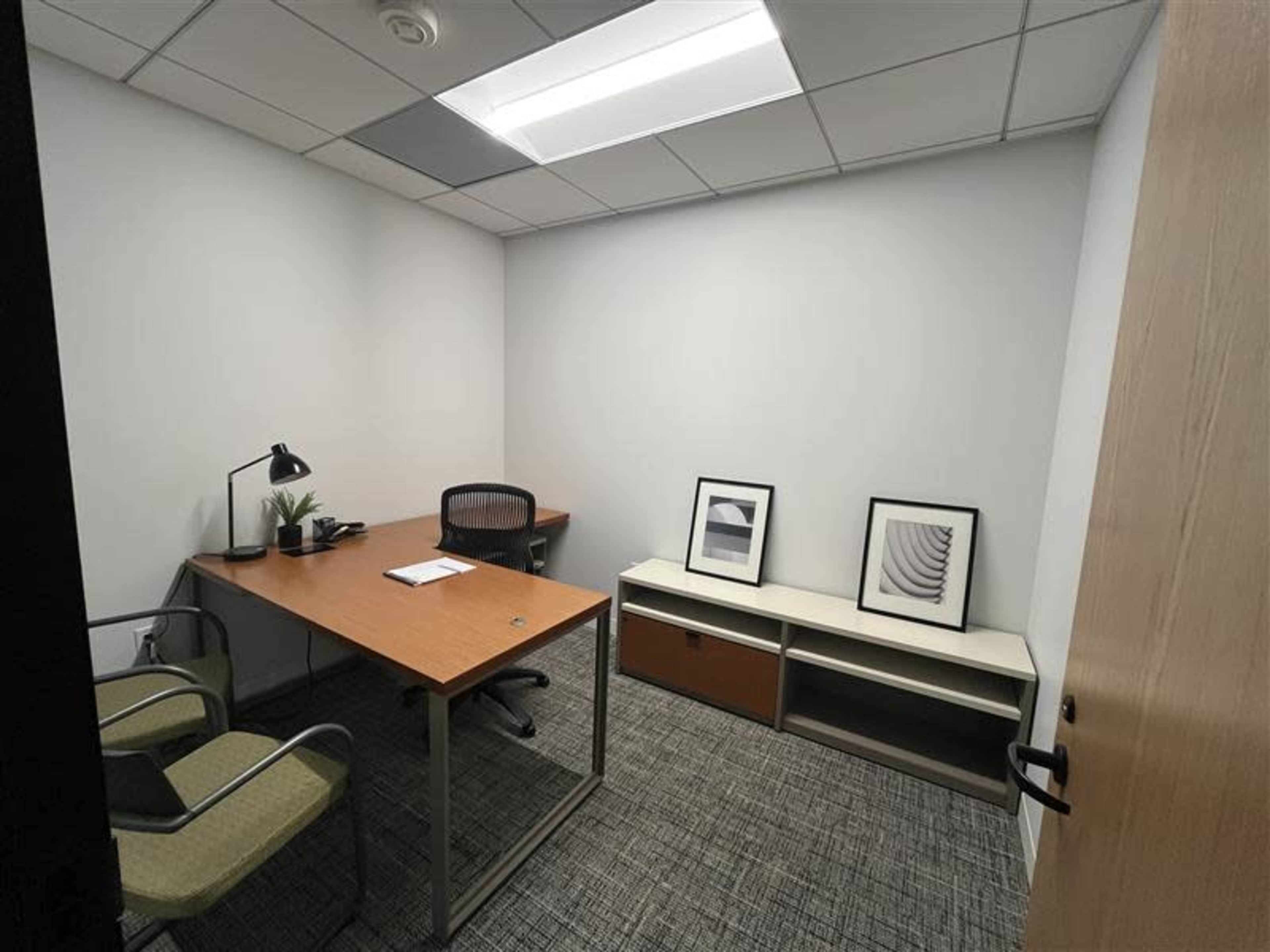 The image shows a small, minimalistic office with a wooden desk, two chairs, and framed artwork on the wall.