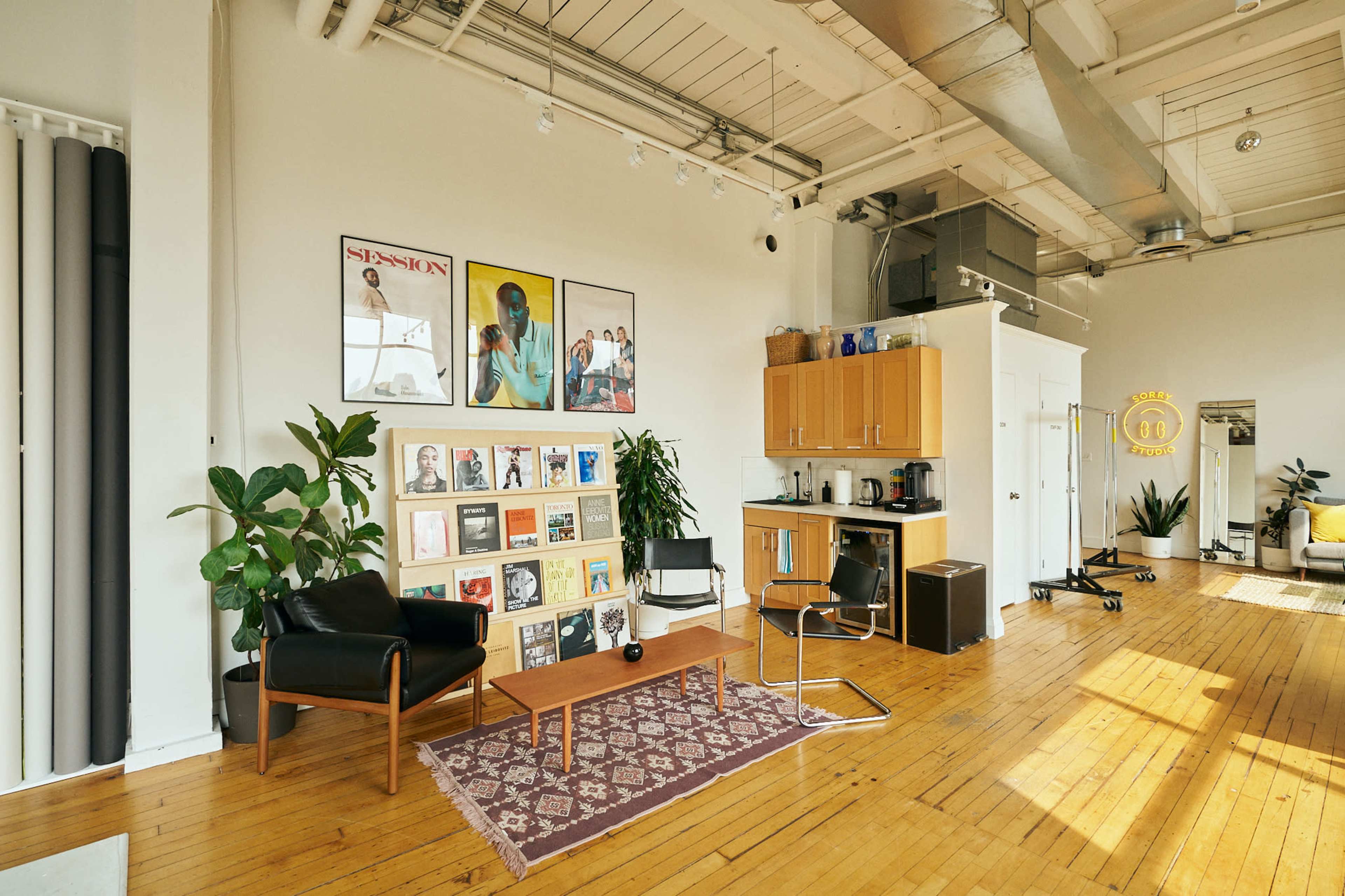 The image shows a spacious, modern interior with a seating area, wooden flooring, artwork on the walls, and a kitchenette along one side.