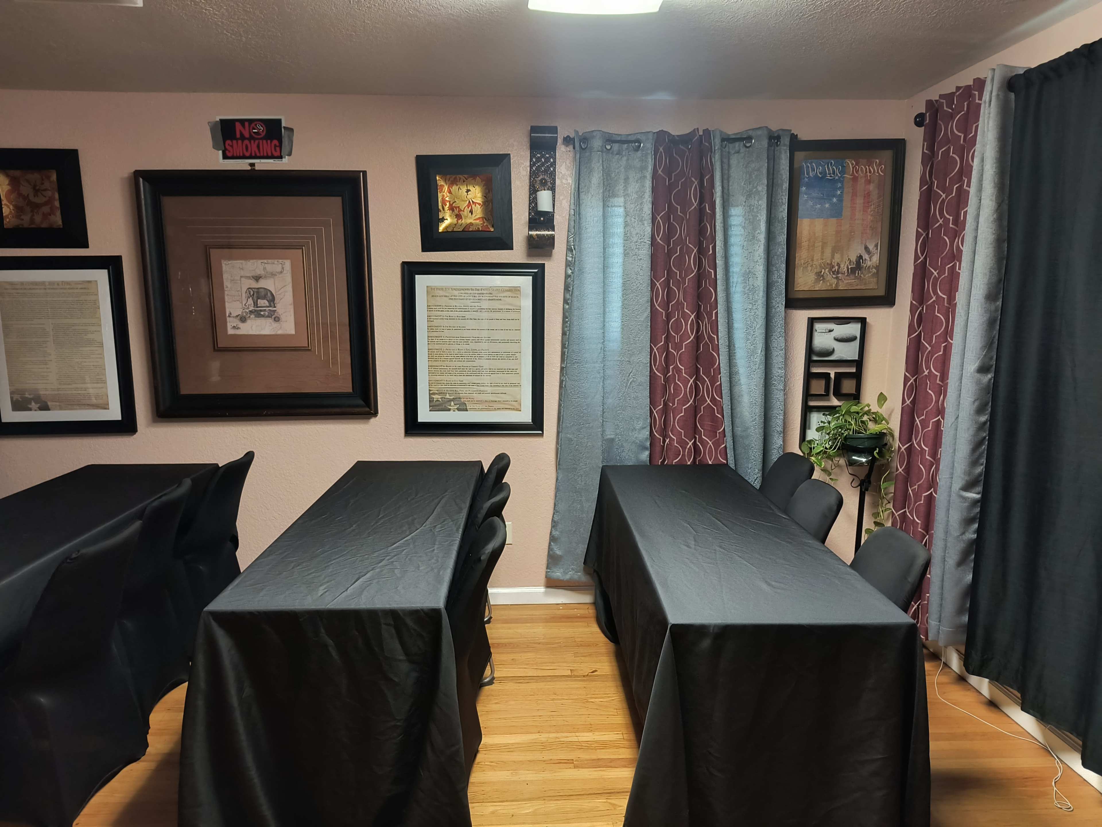 The image shows a room with two tables covered in black cloth, surrounded by framed artwork and curtains on the windows.
