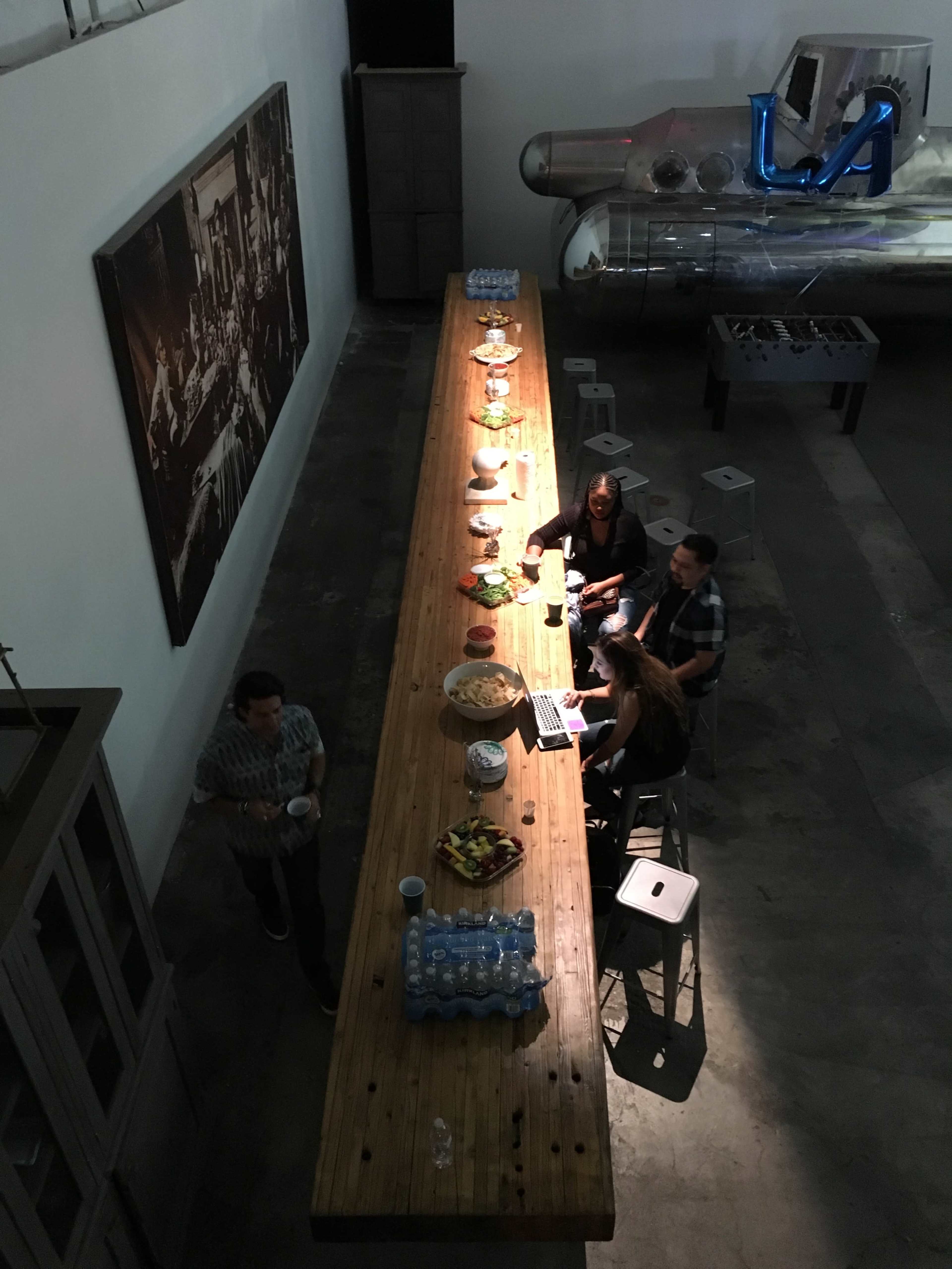 A long wooden table is set with food and drinks, while four people sit around it, and a metallic sculpture is visible in the background.