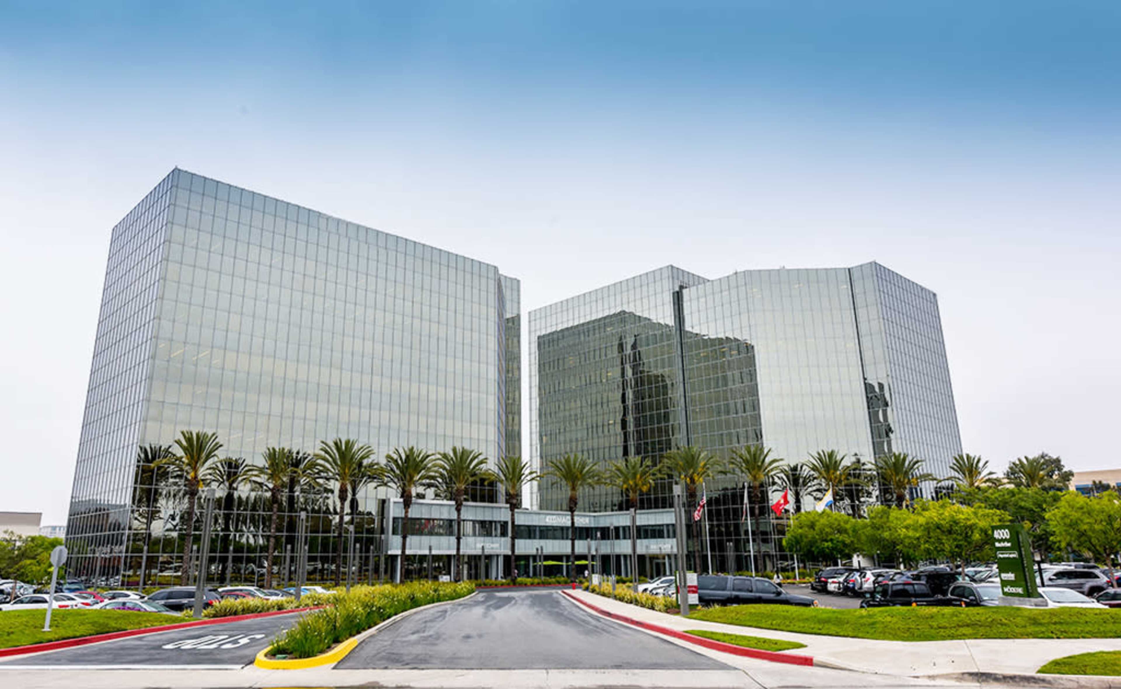 The image shows a modern glass office building with palm trees in the foreground and a parking area in front.