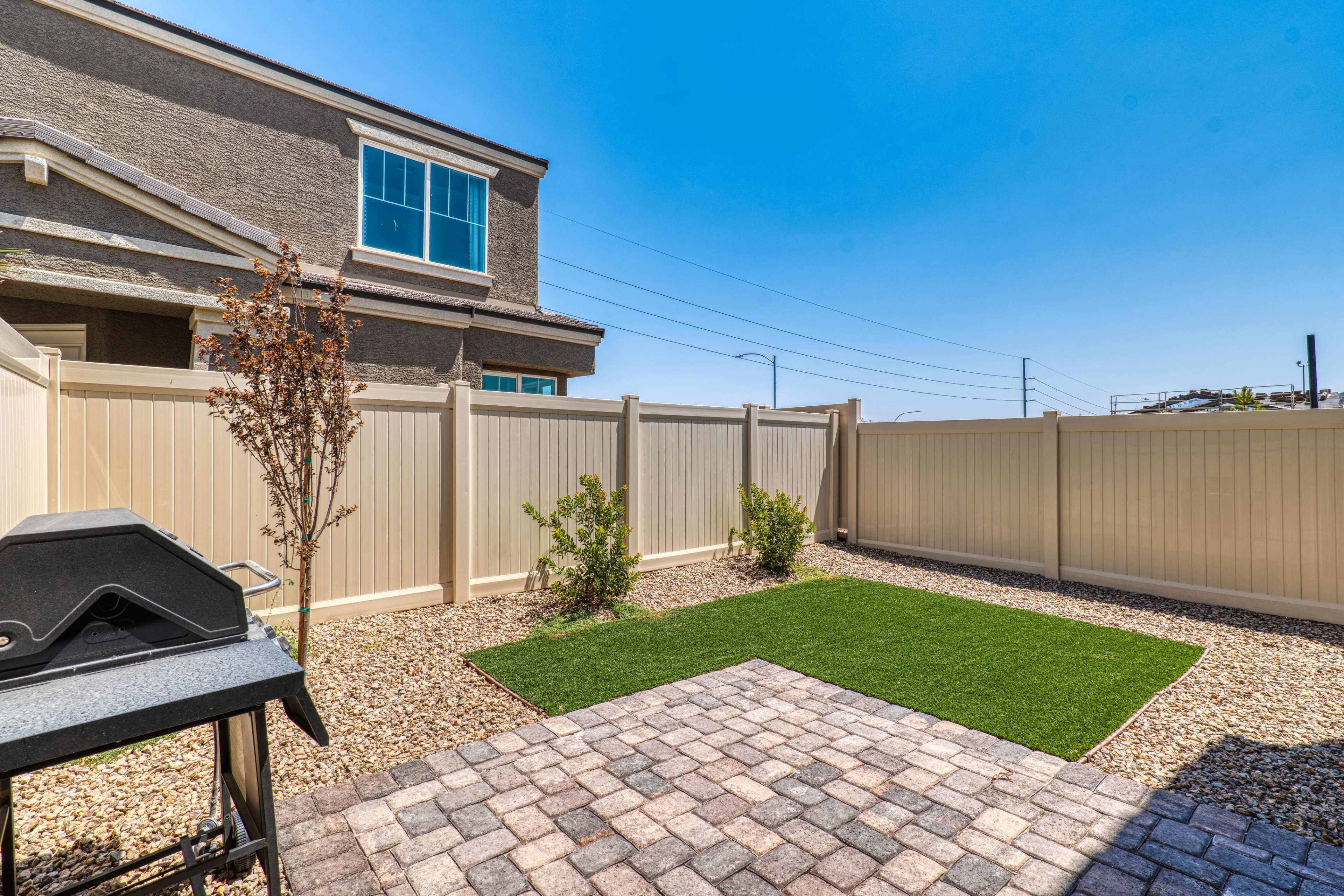 A small backyard with a paved area, artificial grass, and a grill, enclosed by a high fence and adjoining a house.