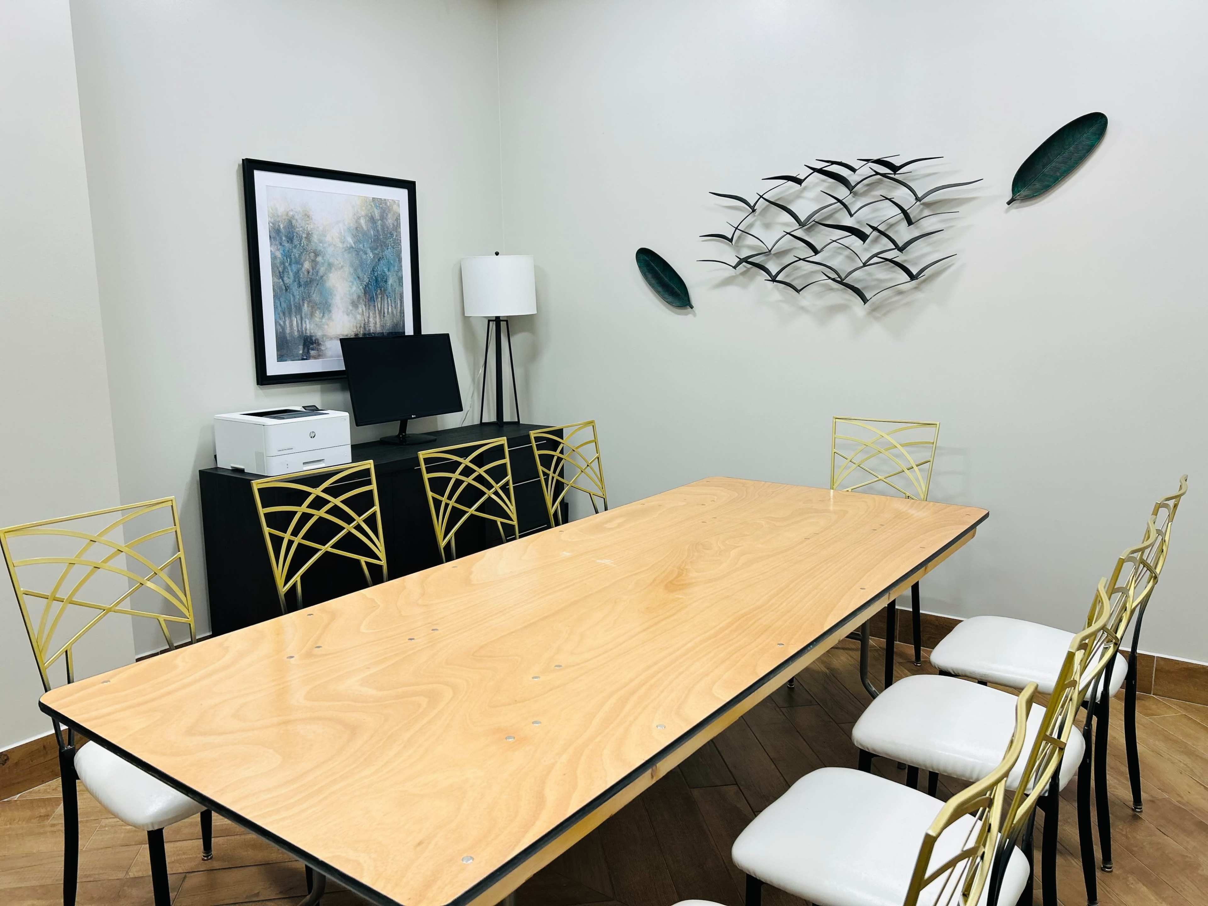 A modern conference room features a wooden table surrounded by six chairs, a wall-mounted artwork, a computer monitor on a cabinet, and a decorative piece of birds on the wall.